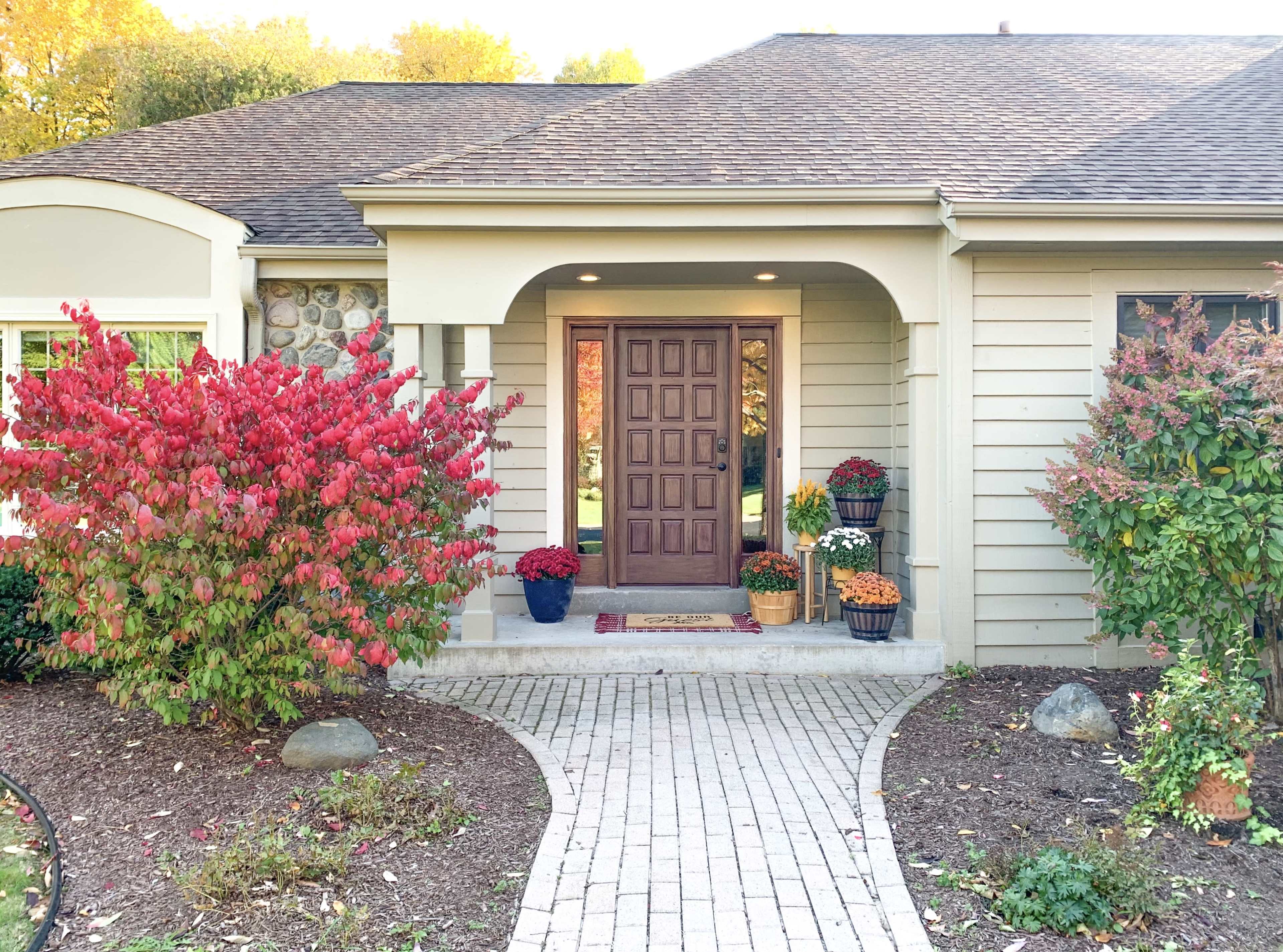 The image shows a front entrance of a house with a brown door, flanked by colorful potted plants and a brick walkway surrounded by blooming shrubs.