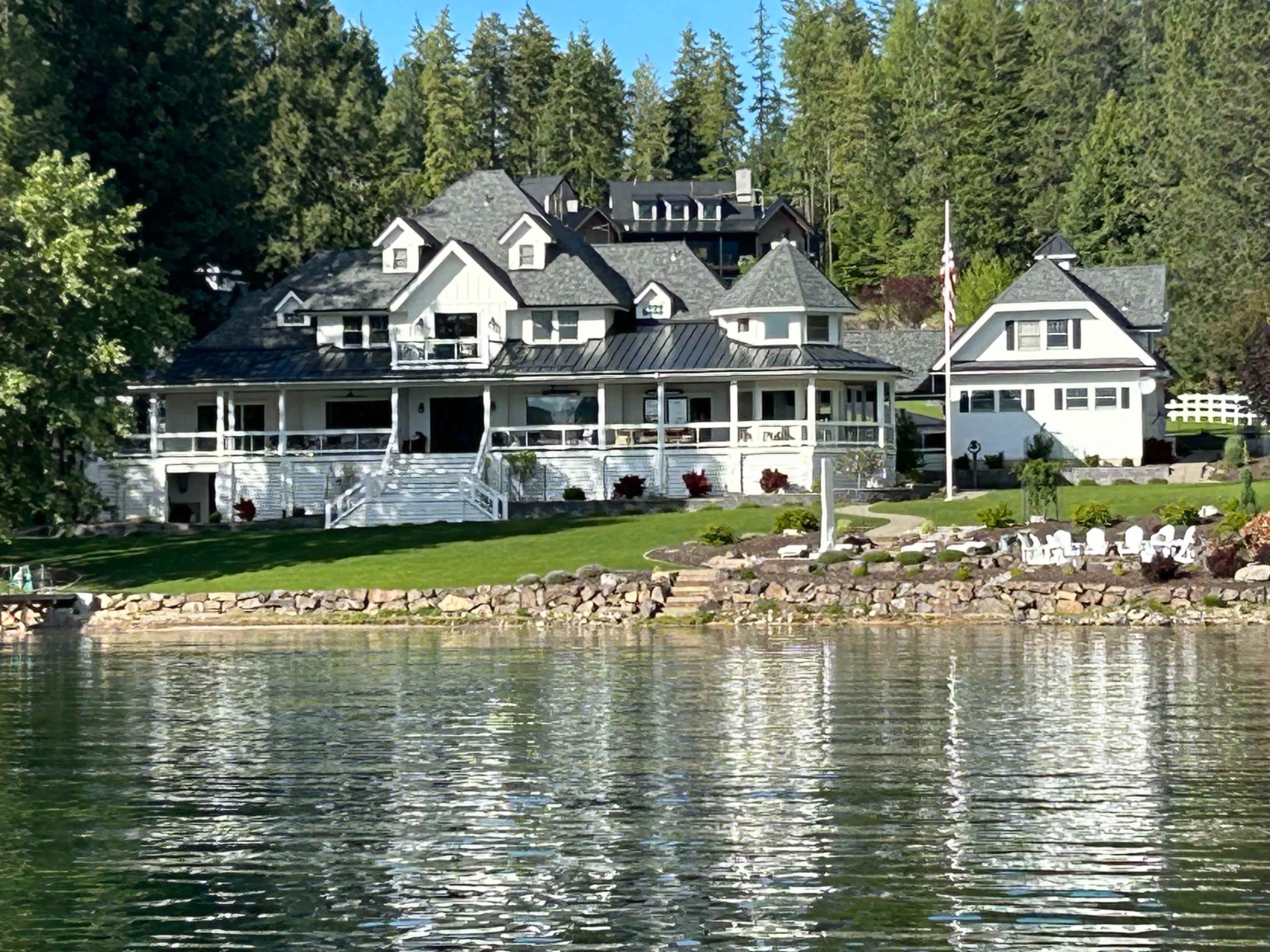 A large, multi-level white house with a wraparound porch and several smaller buildings on a shoreline, surrounded by trees and reflecting in calm water.