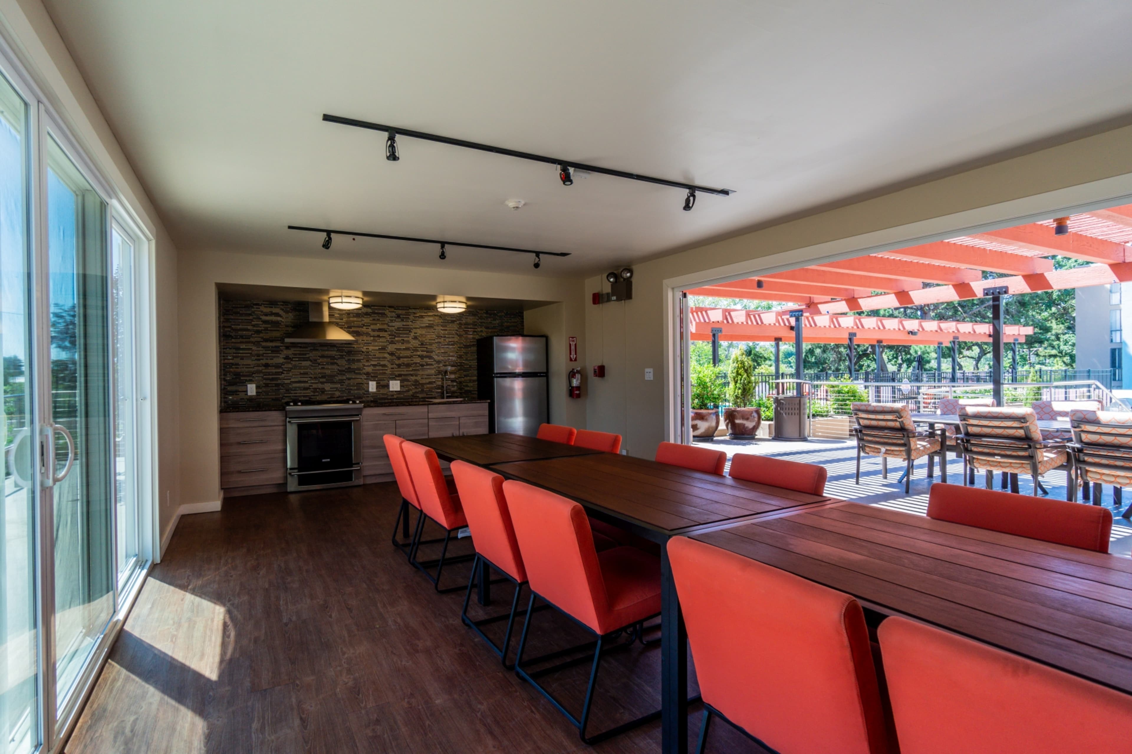 The image shows a modern indoor kitchen and dining area with a long table and orange chairs, leading to an outdoor space with seating under a pergola.