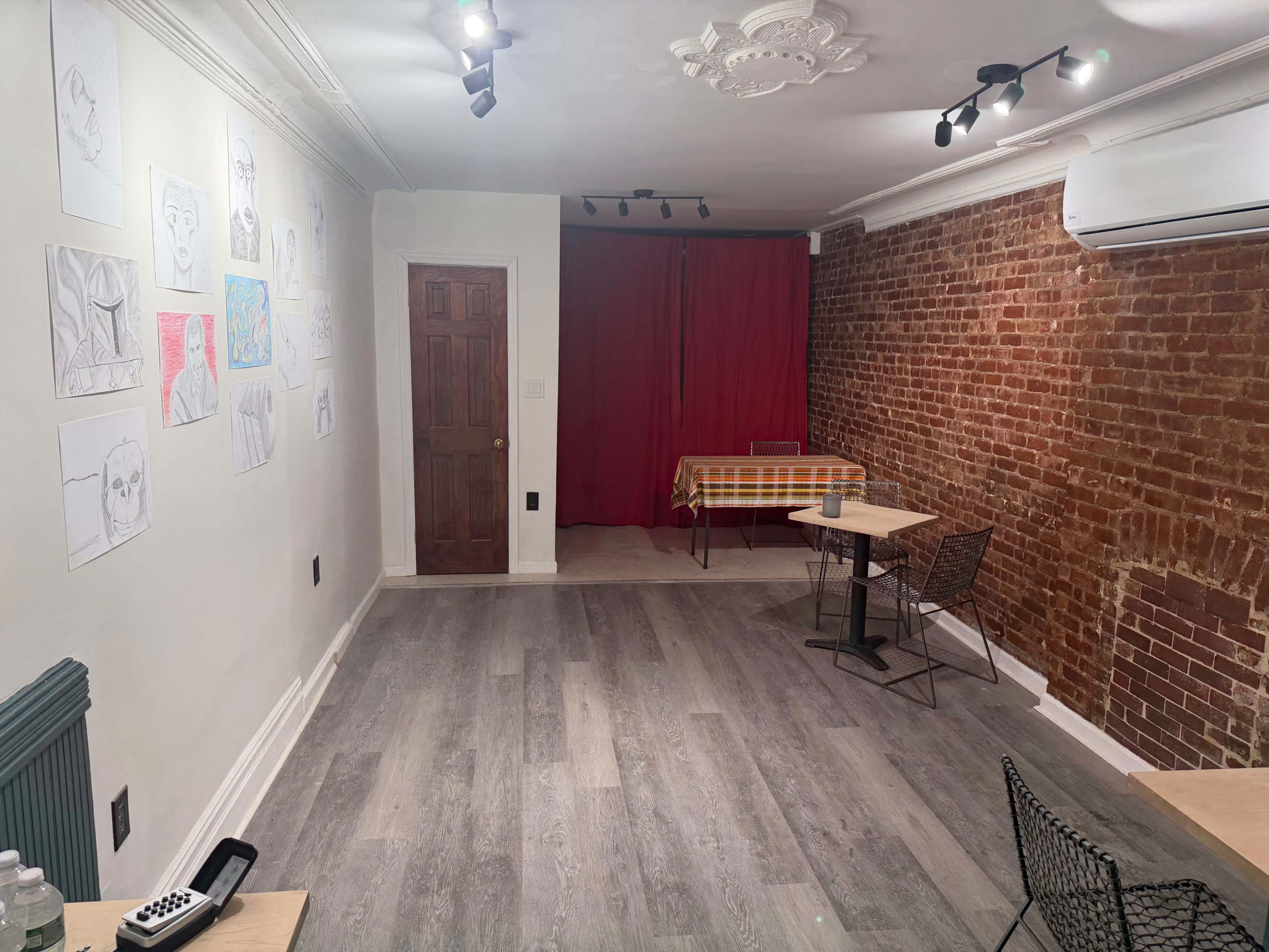 The image shows a room with exposed brick walls, featuring a table with a checkered tablecloth, two small tables with chairs, and a series of drawings hung on the wall.