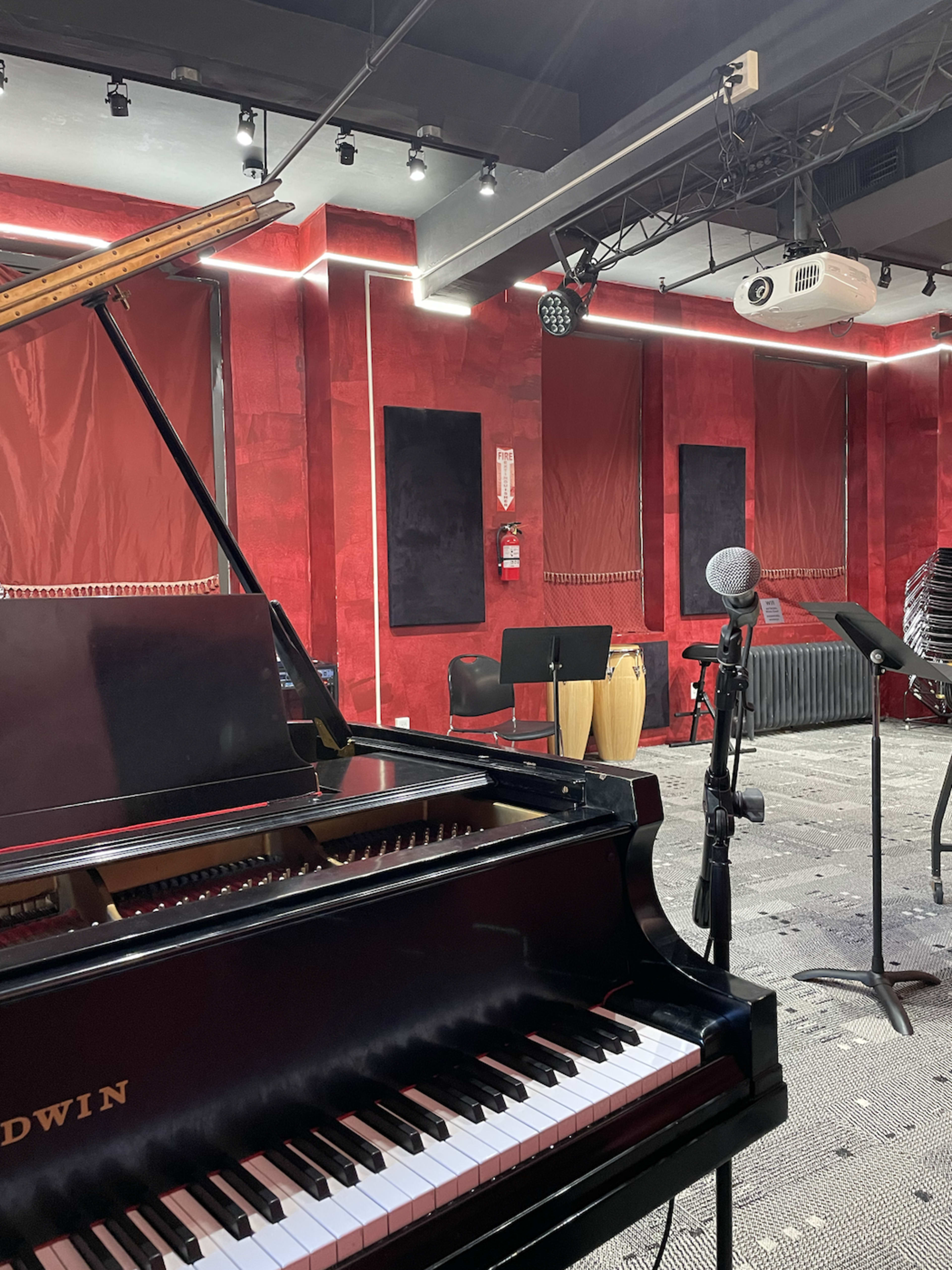 A black grand piano in a music studio with red walls, a microphone on a stand, and various instruments, including congas.