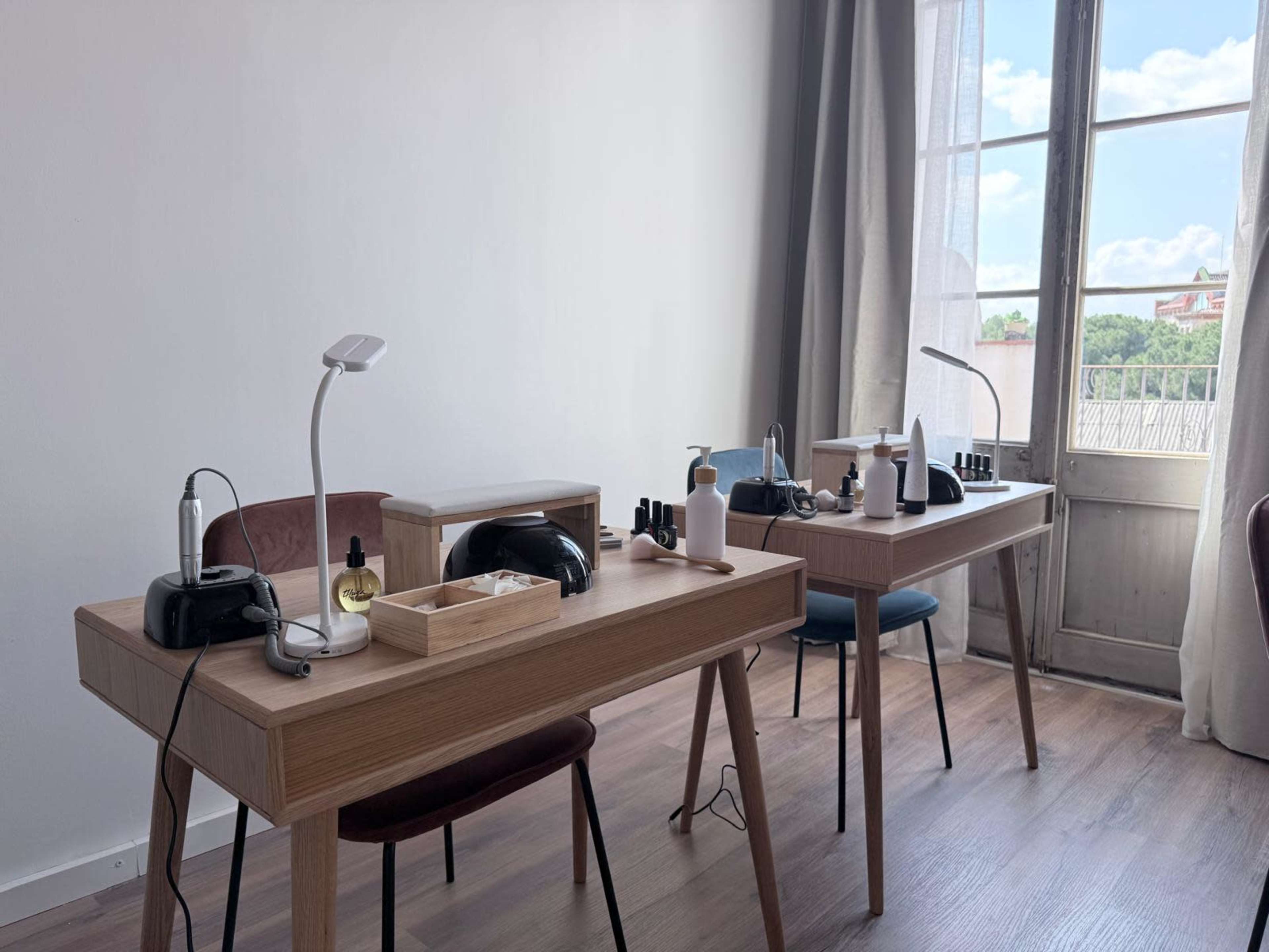 The image shows two wooden manicure tables set up in a well-lit room with a window, tools, and products for nail care arranged neatly on each table.