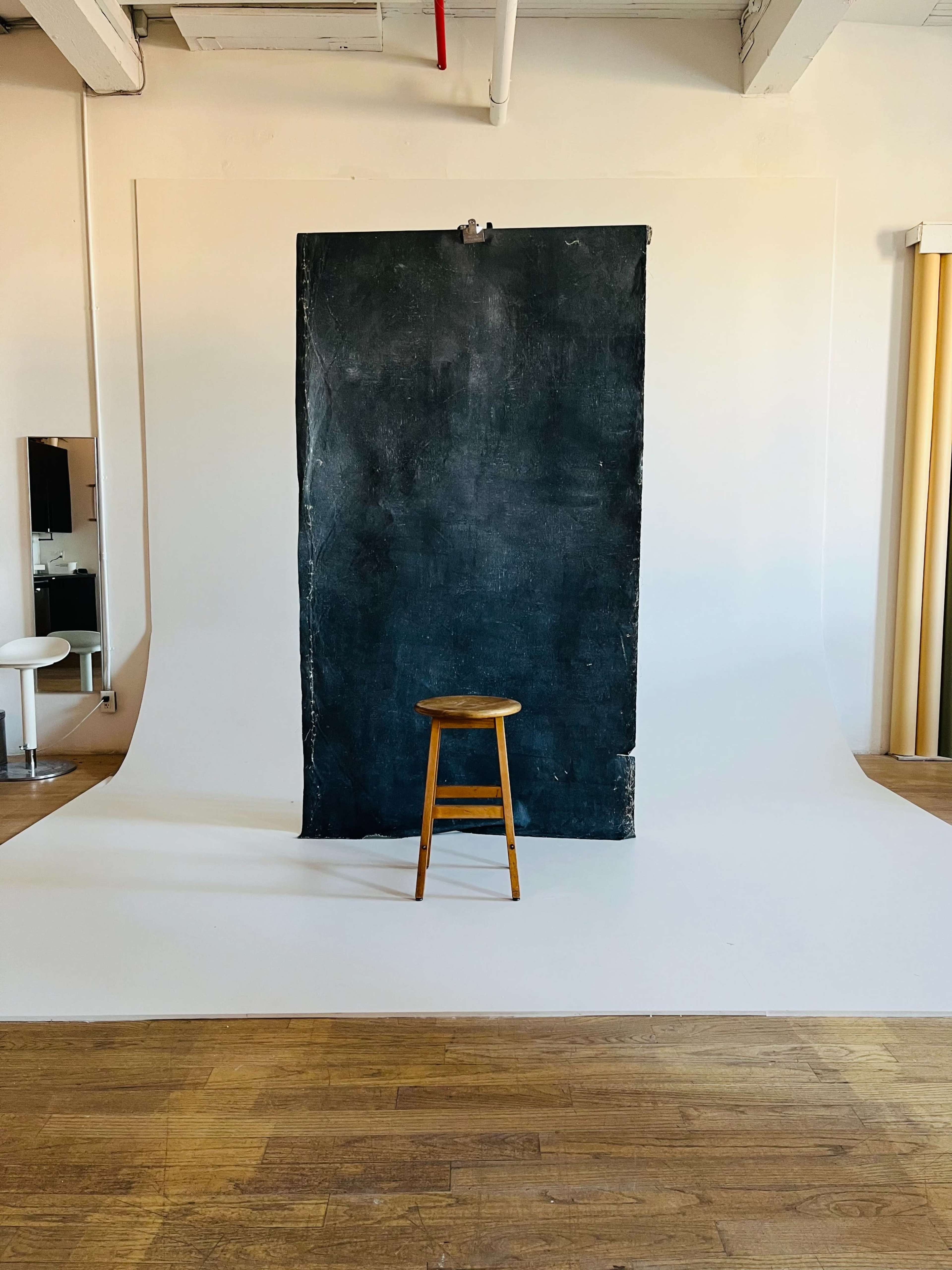A wooden stool stands in front of a large, dark backdrop within a photo studio.