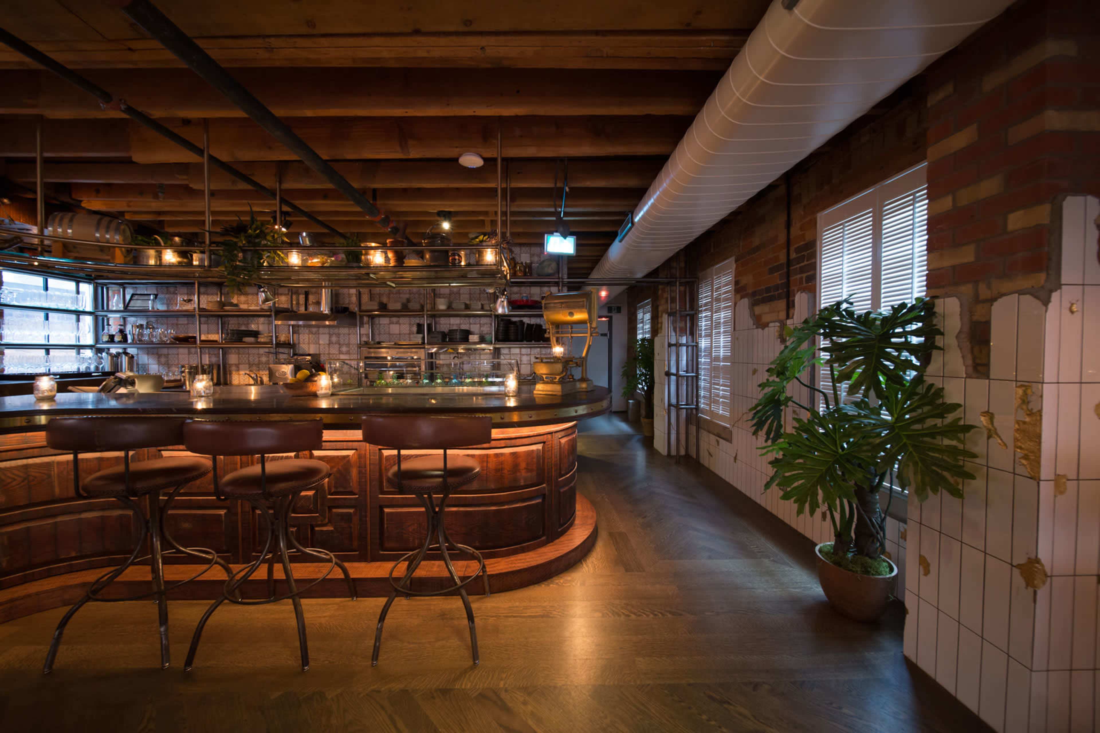 The image shows a modern bar area with wooden counter stools, a well-lit countertop, and a stylish interior featuring exposed brick and wooden beams.