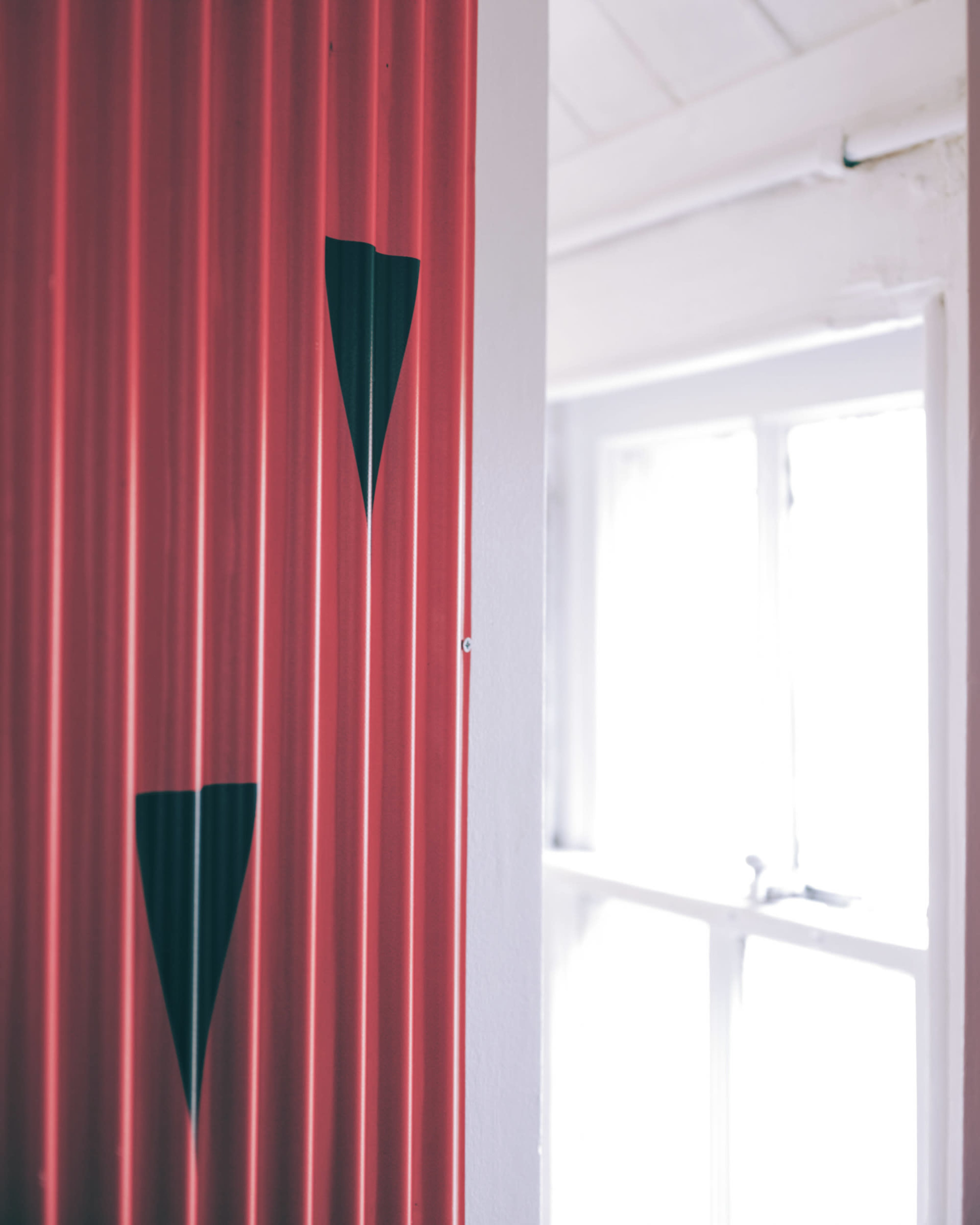 The image shows a section of a wall with red corrugated material featuring two black triangular shapes, alongside a white wall and a window in the background.