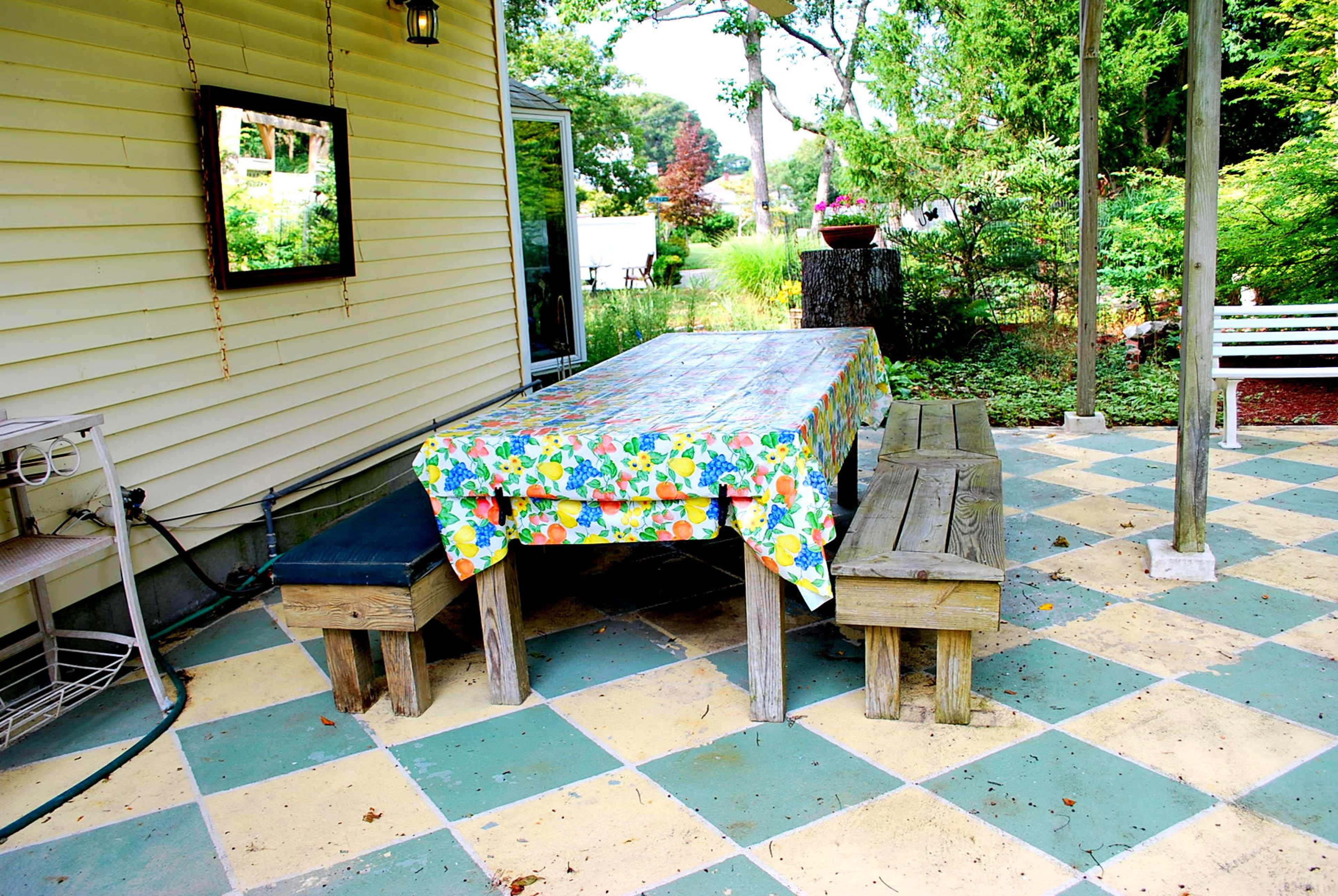 A wooden table covered with a colorful tablecloth is set in a garden area alongside two wooden benches and a patio featuring a checkerboard pattern.