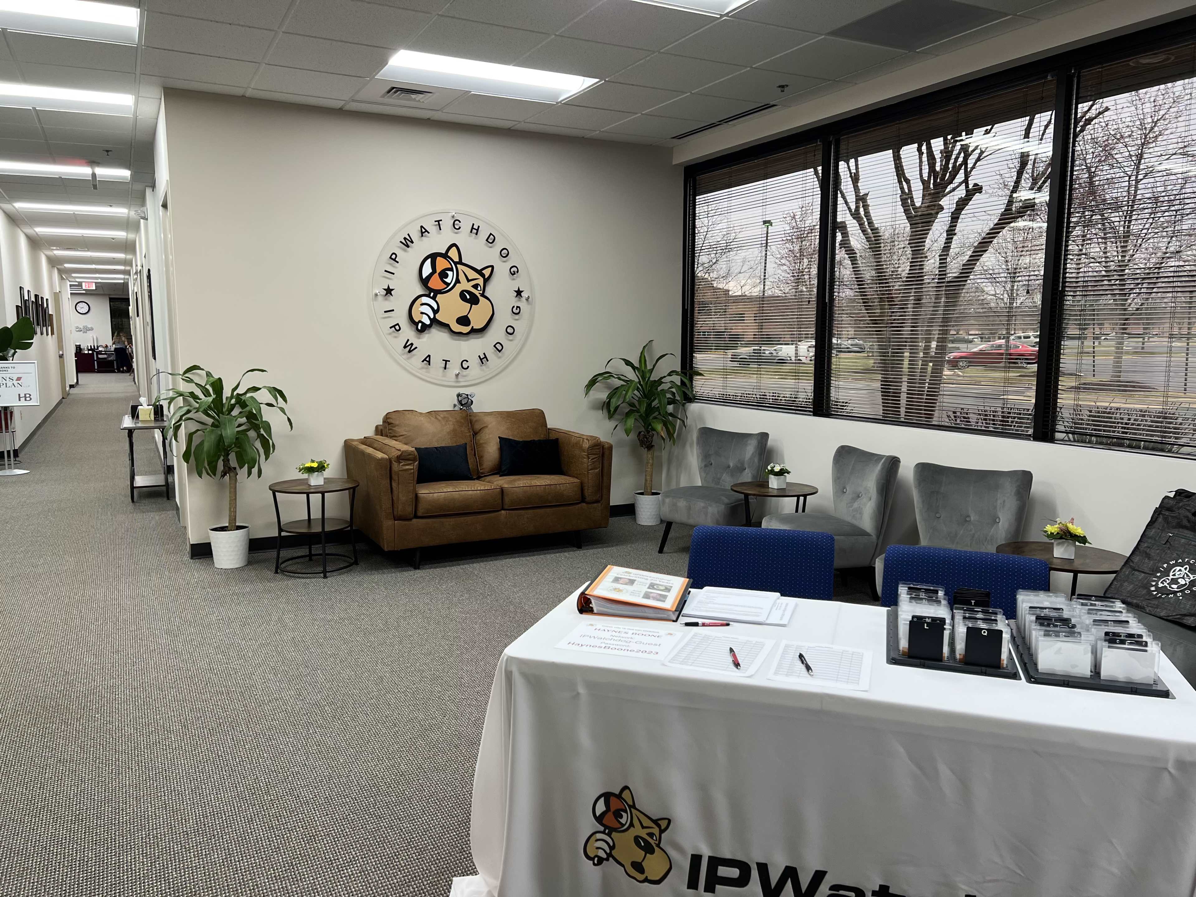 A modern office hallway with a reception area featuring a couch, plants, and a table displaying informational materials and sign-in sheets.