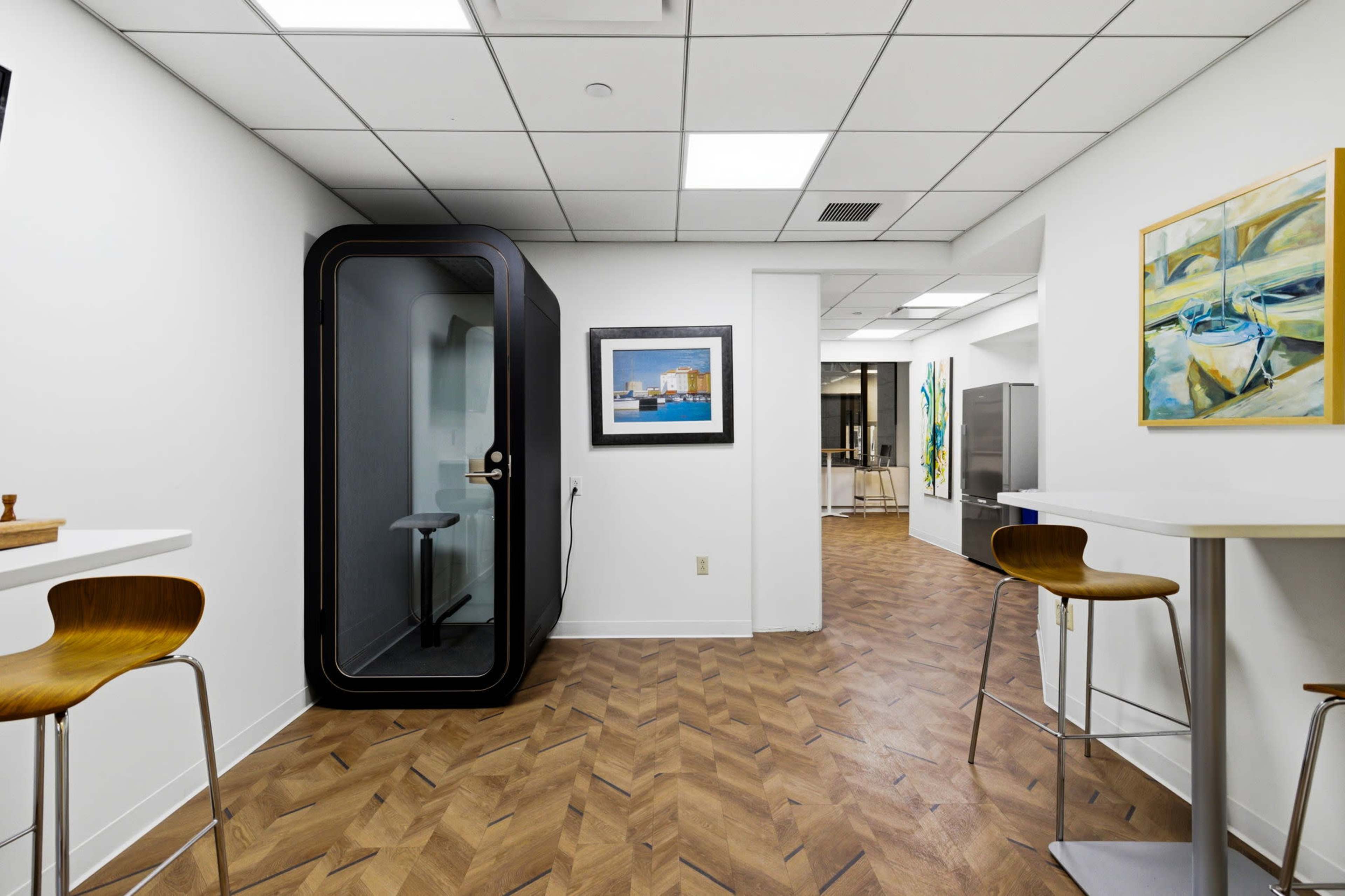 The image shows a modern office space with a phone booth in the corner, high stools at small tables, and artwork on the walls.