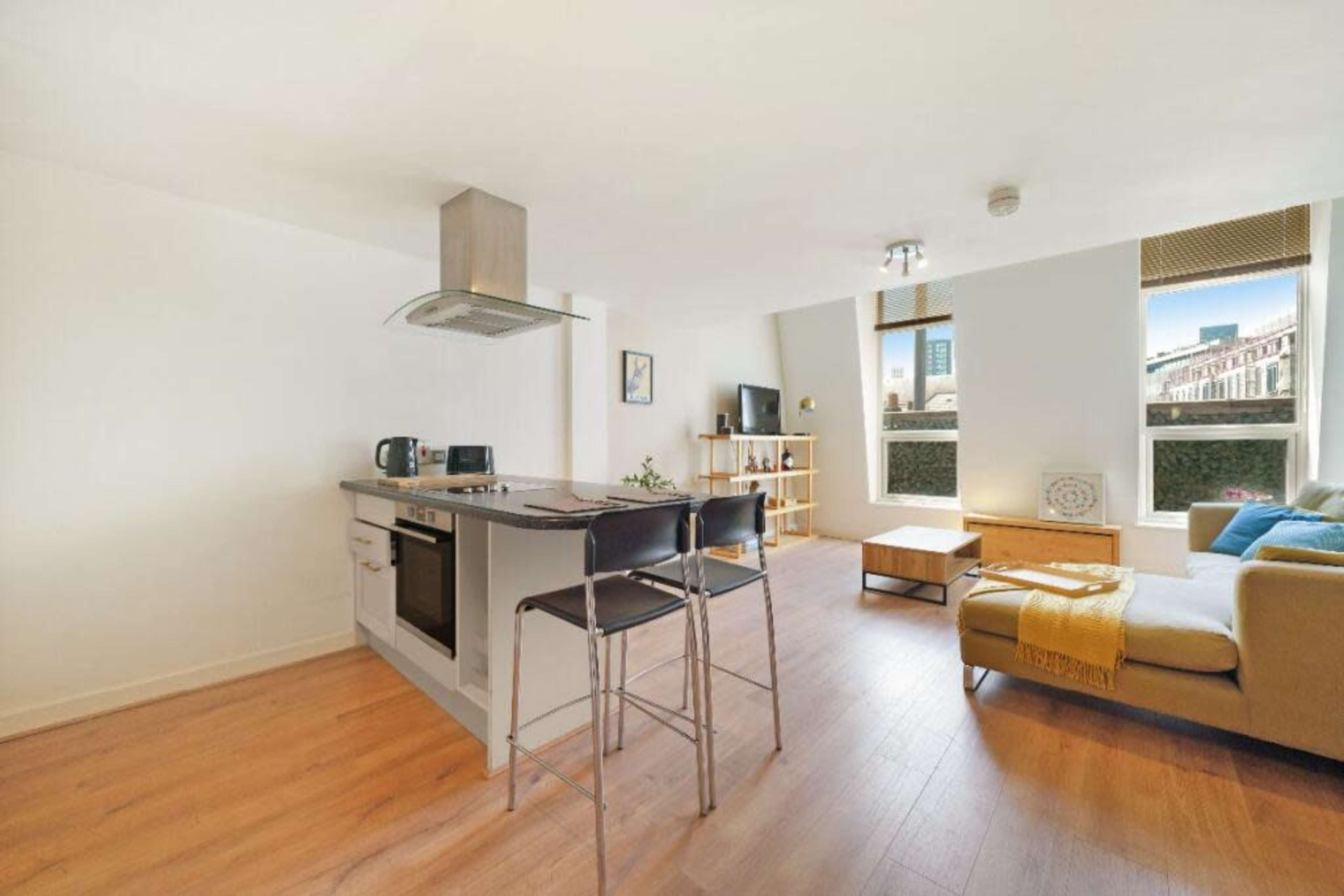 The image depicts a modern, open-concept living space featuring a kitchen island with seating, a sofa, and large windows with urban views.