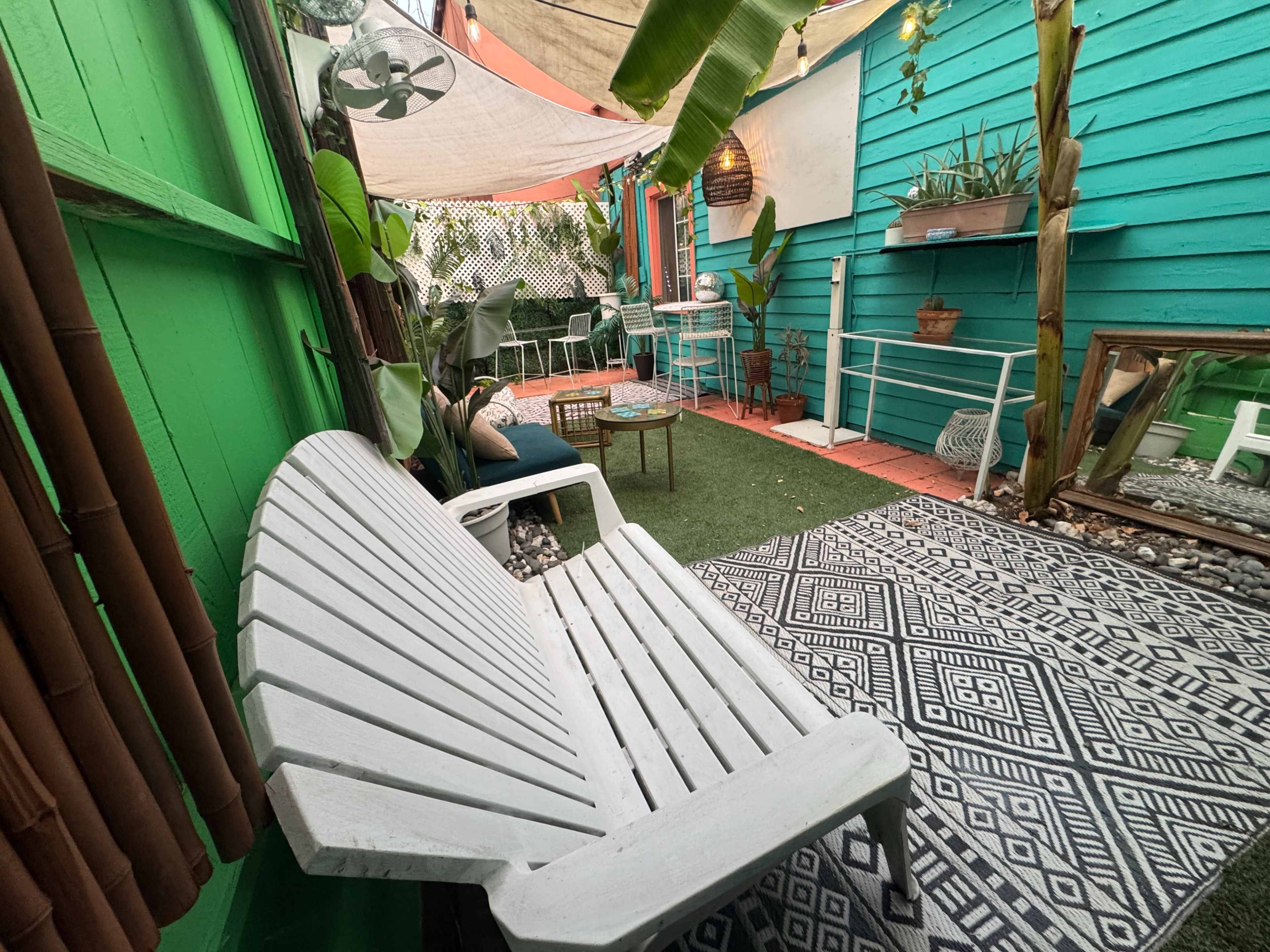 The image shows a small outdoor garden area with a white lounge chair, a patterned rug, and various plants against colorful walls.