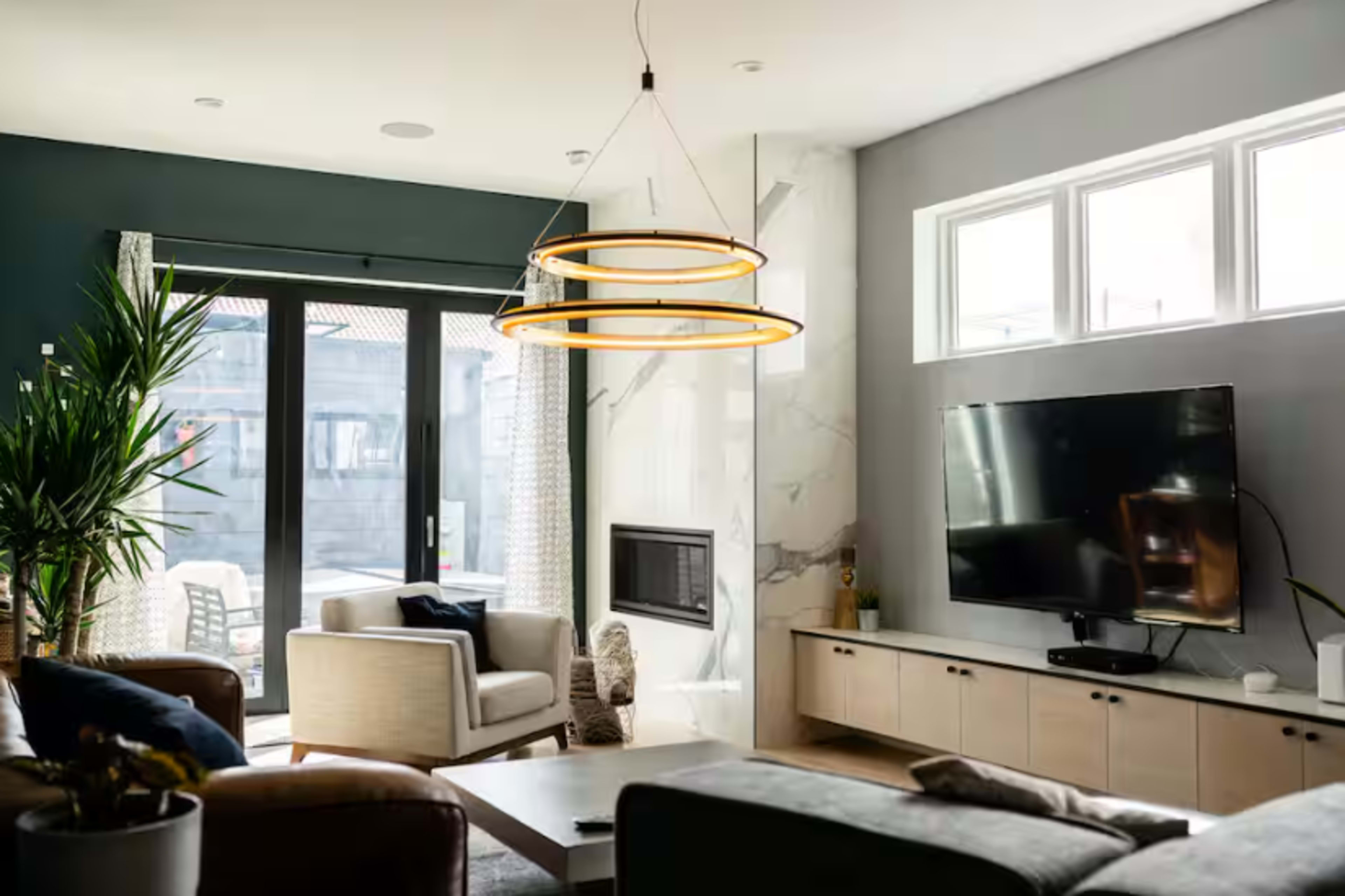 The living room features a modern design with a large television, a minimalist entertainment unit, and a two-tiered pendant light hanging from the ceiling, surrounded by large windows and indoor plants.