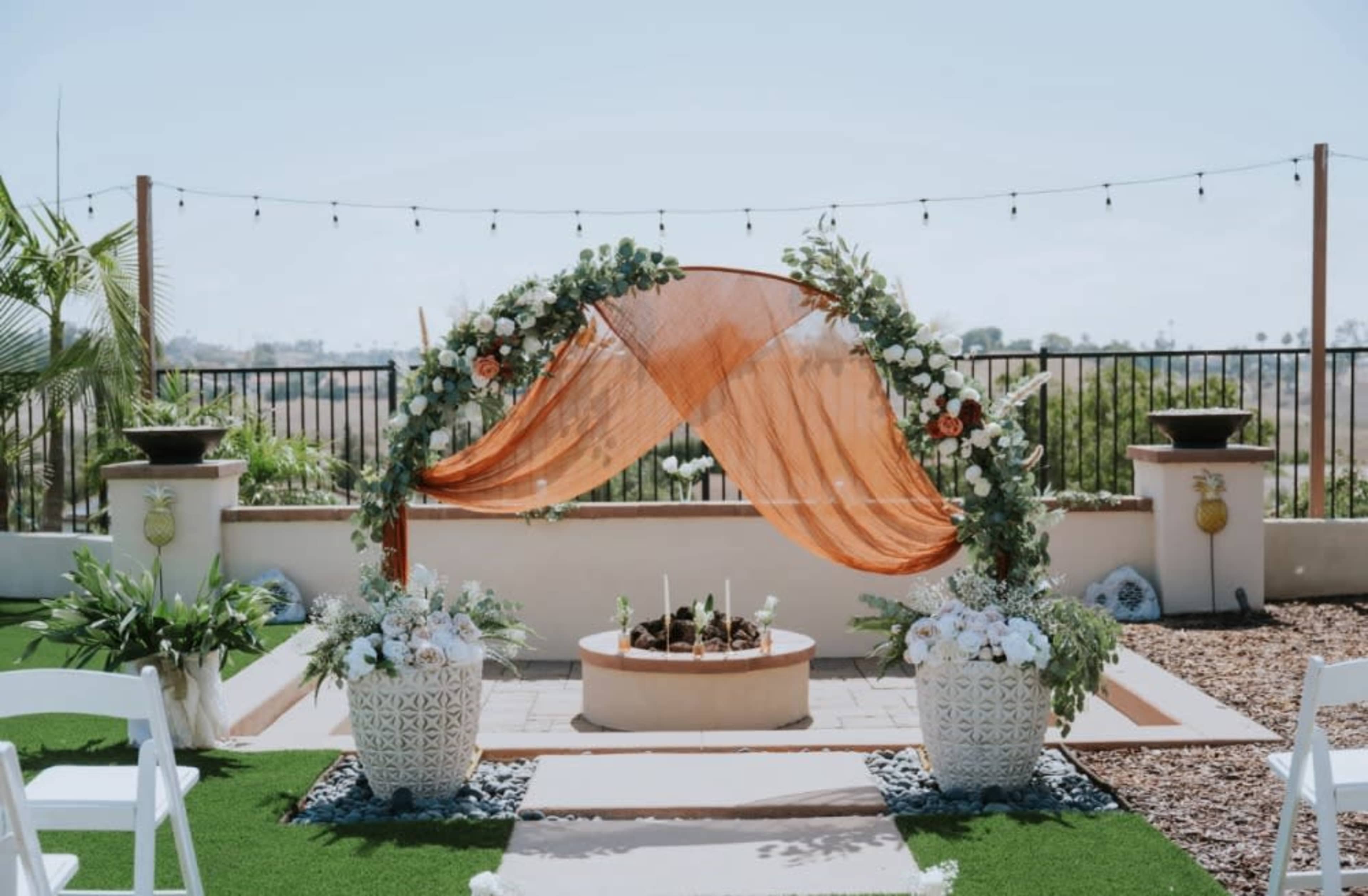 The image features a wedding altar adorned with white flowers and an orange draped fabric, set in a landscaped outdoor area with potted plants.