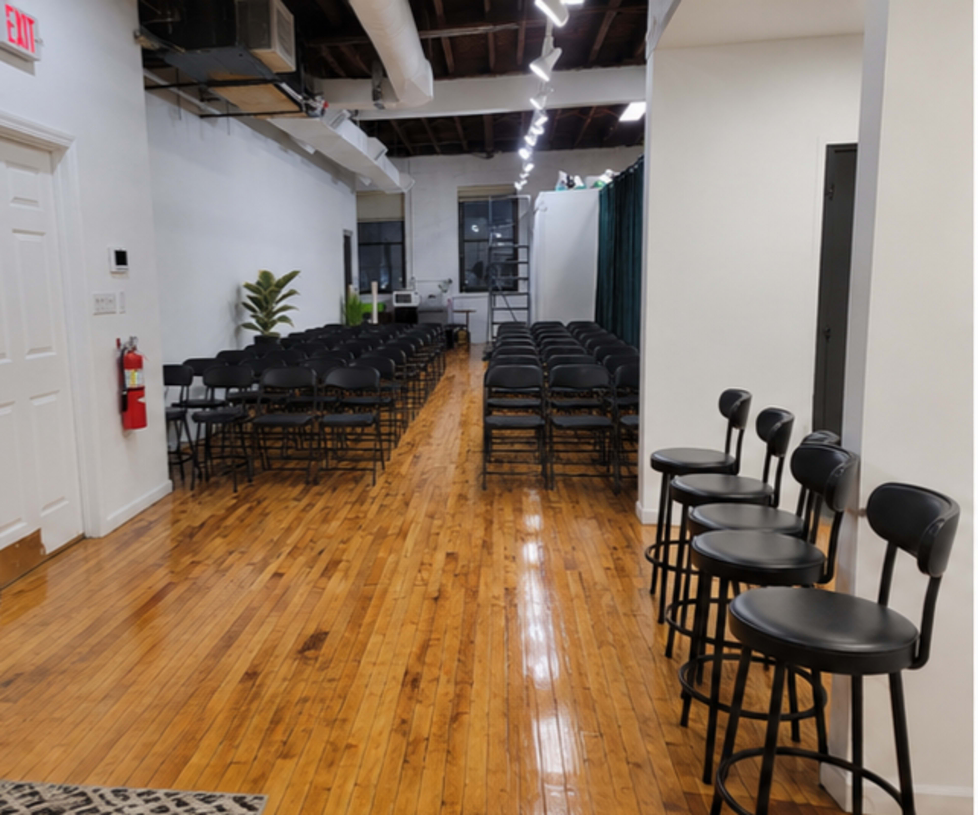 The image shows a narrow, well-lit room with a row of folding chairs set up in the center and several tall bar stools lined up against one side.