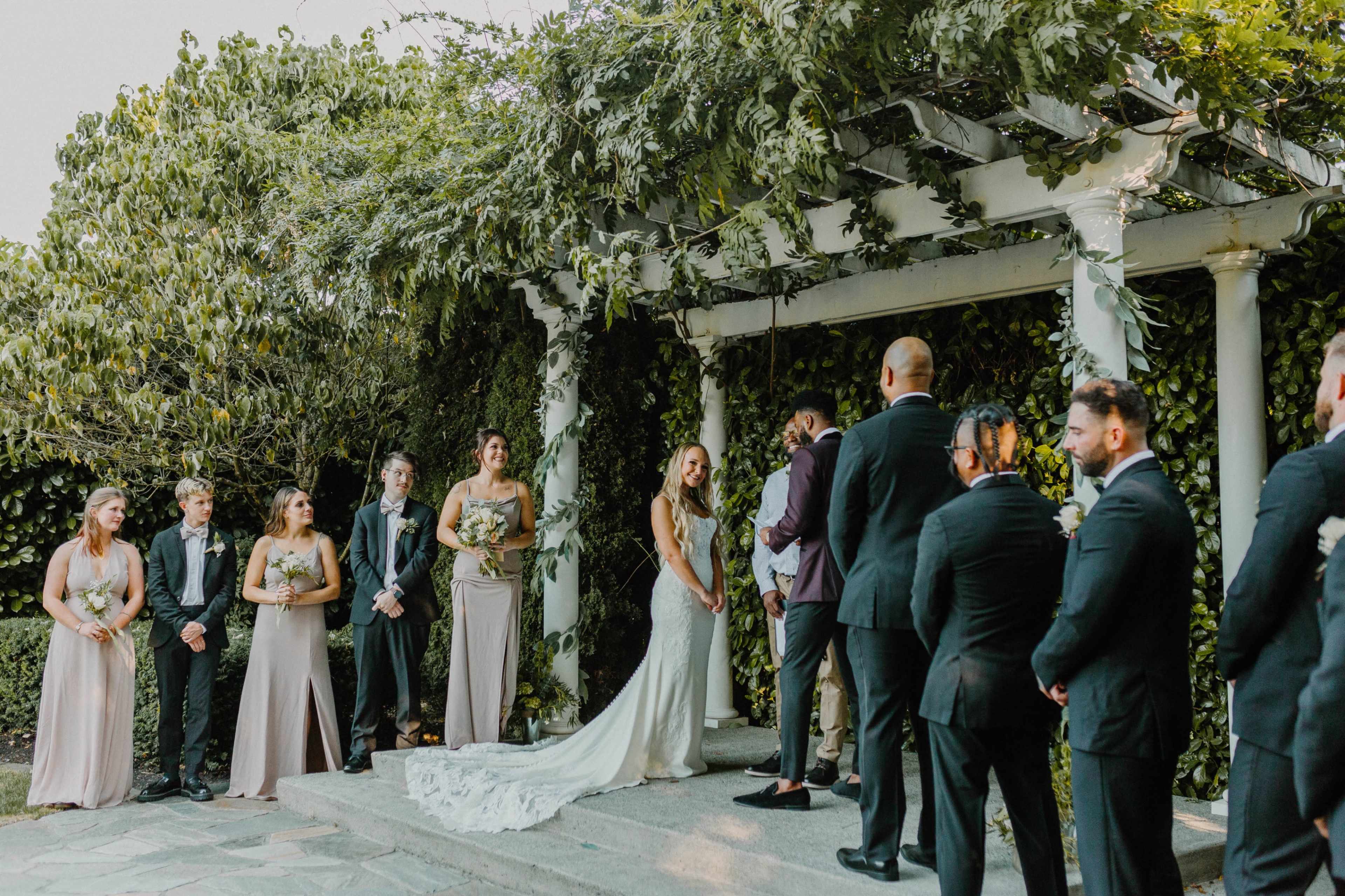 A couple stands under a decorated gazebo exchanging vows, surrounded by their wedding party and guests.