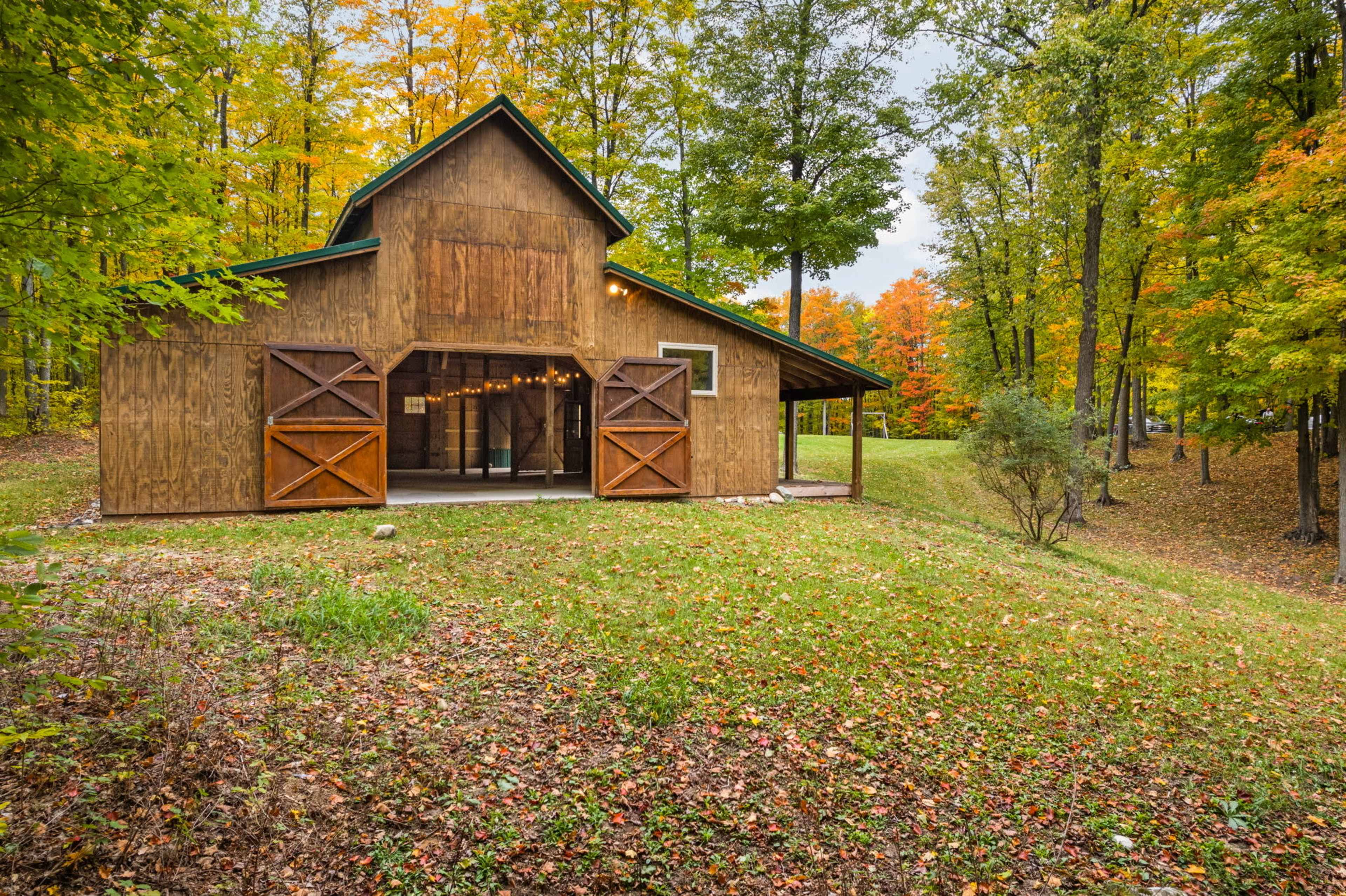 A wooden barn with large doors stands in a grassy area surrounded by trees with autumn foliage.