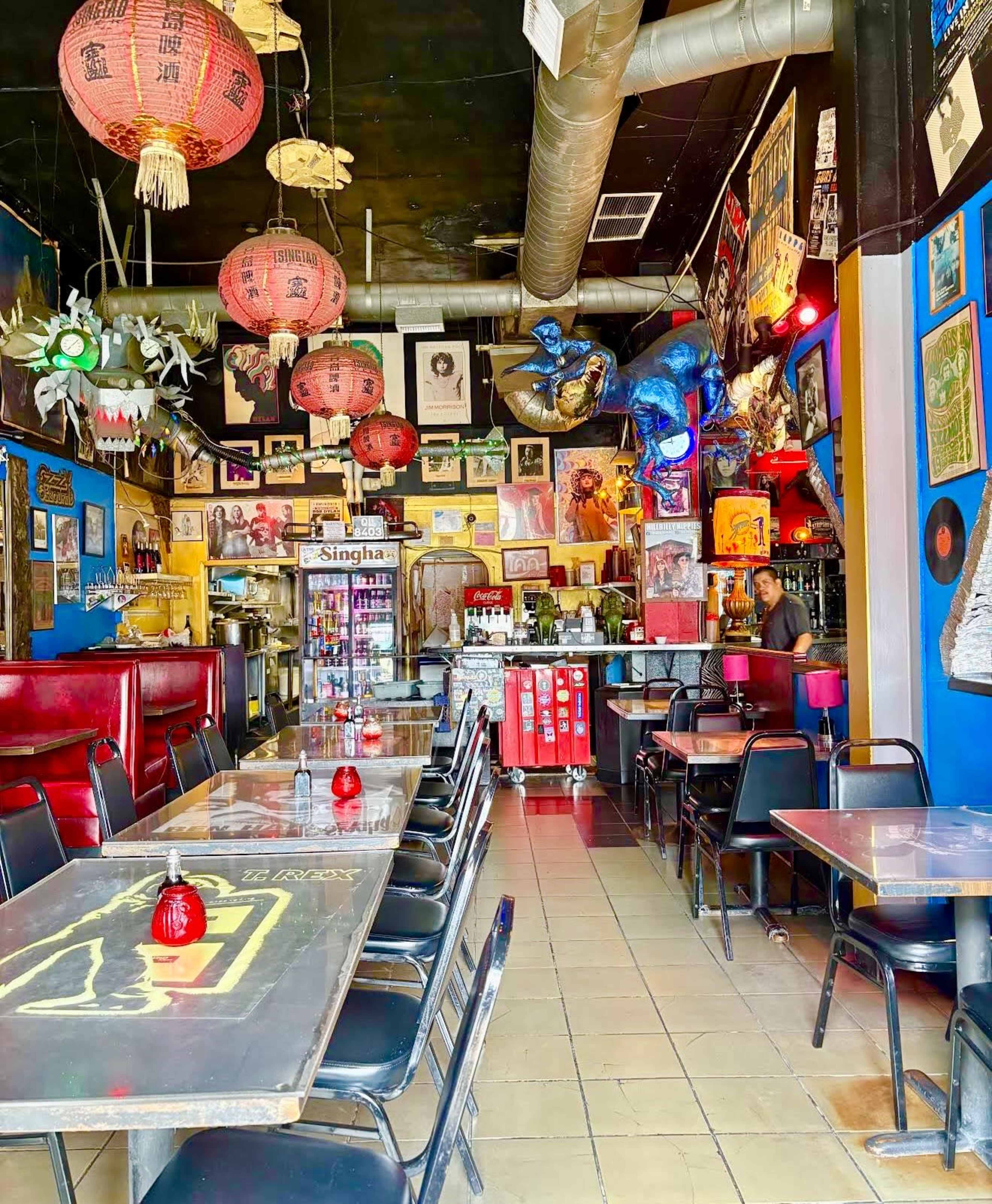 The image shows the interior of a vibrant restaurant, featuring colorful walls adorned with various artworks and decorations, and several tables arranged in a line with red lanterns hanging from the ceiling.