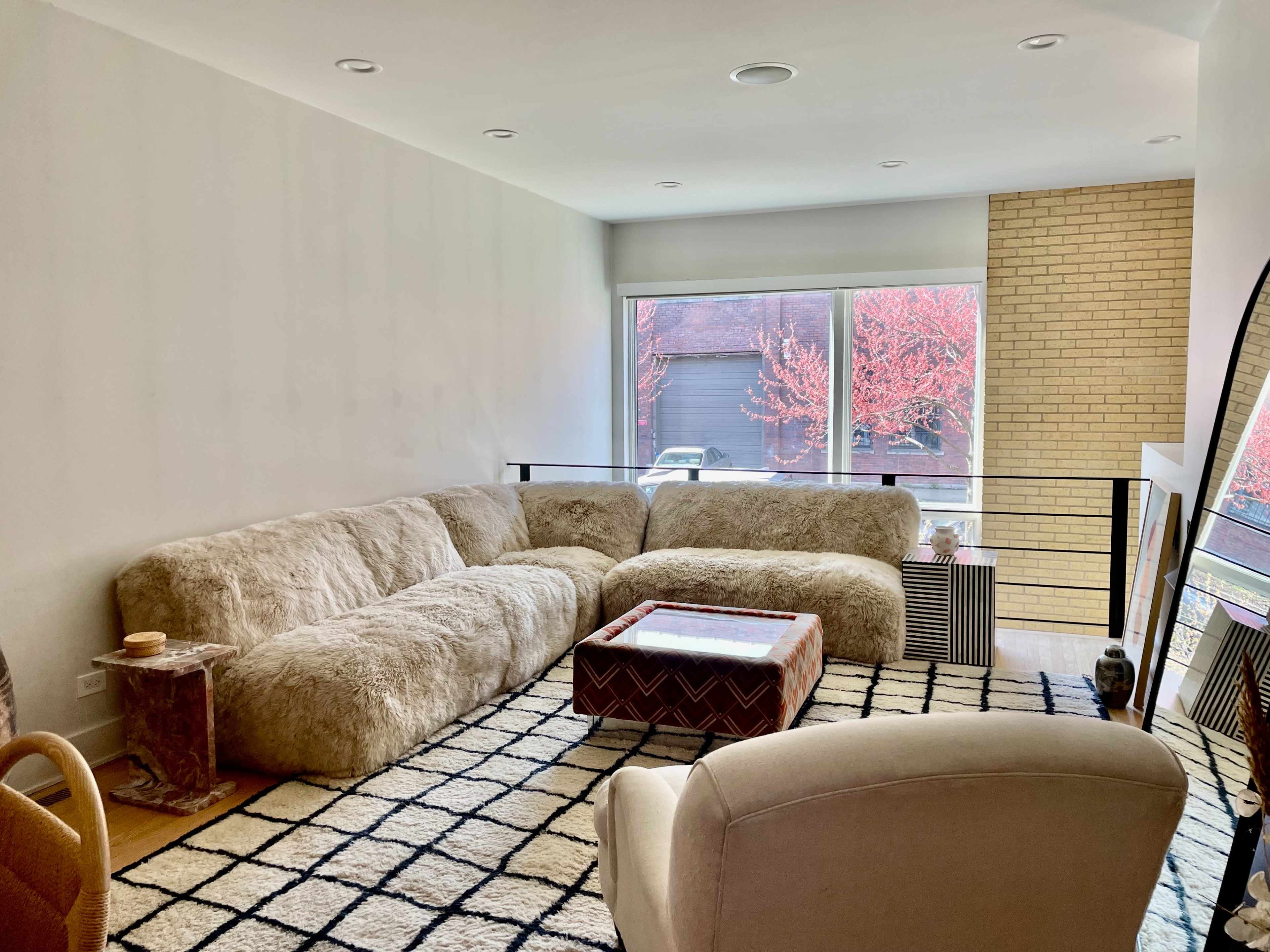 The image features a modern living room with a large, plush sectional sofa, a coffee table, and a view of a vibrant tree outside through large windows.