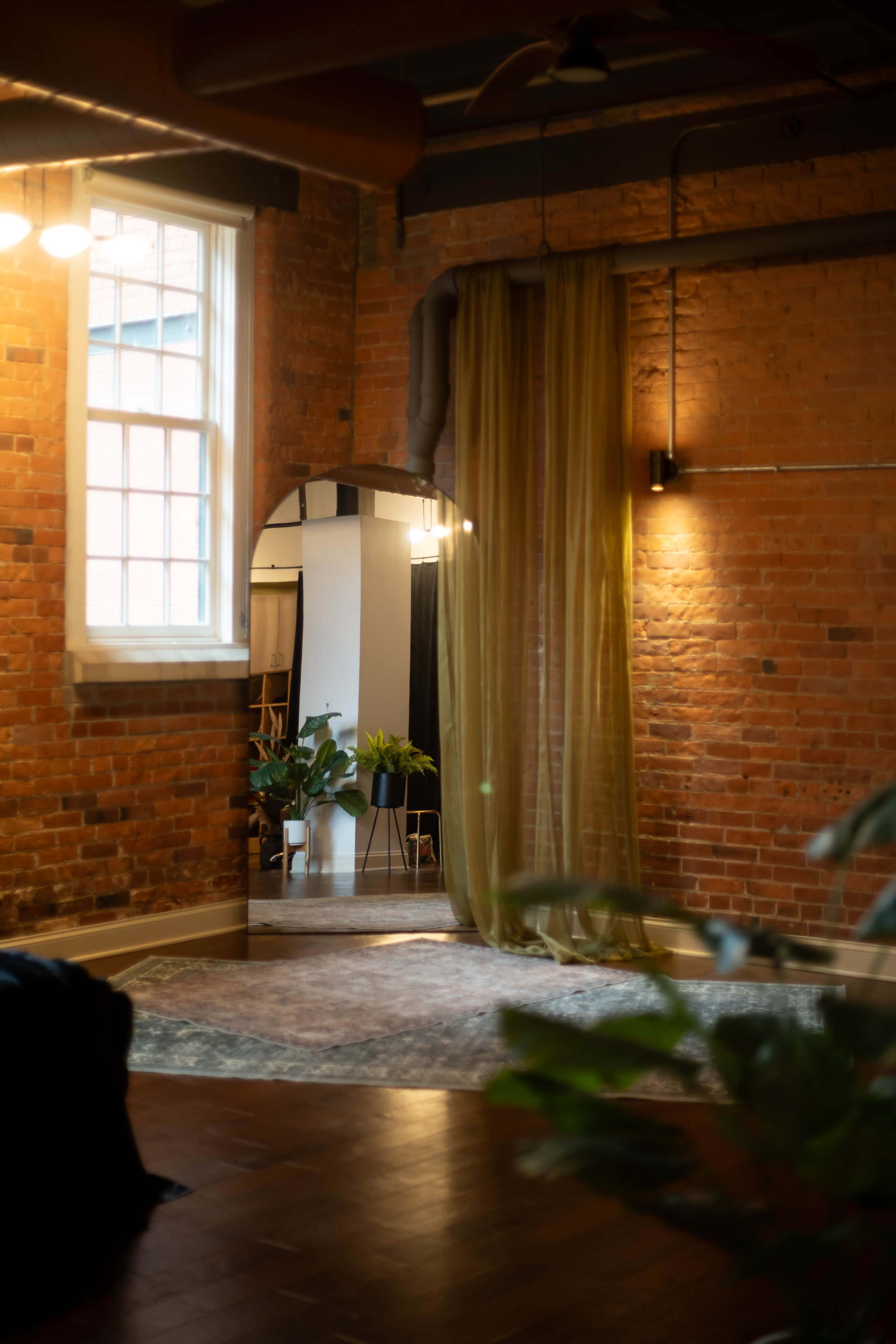 The image shows a room with exposed brick walls, a large mirror, a soft curtain, and plants, creating a cozy atmosphere.