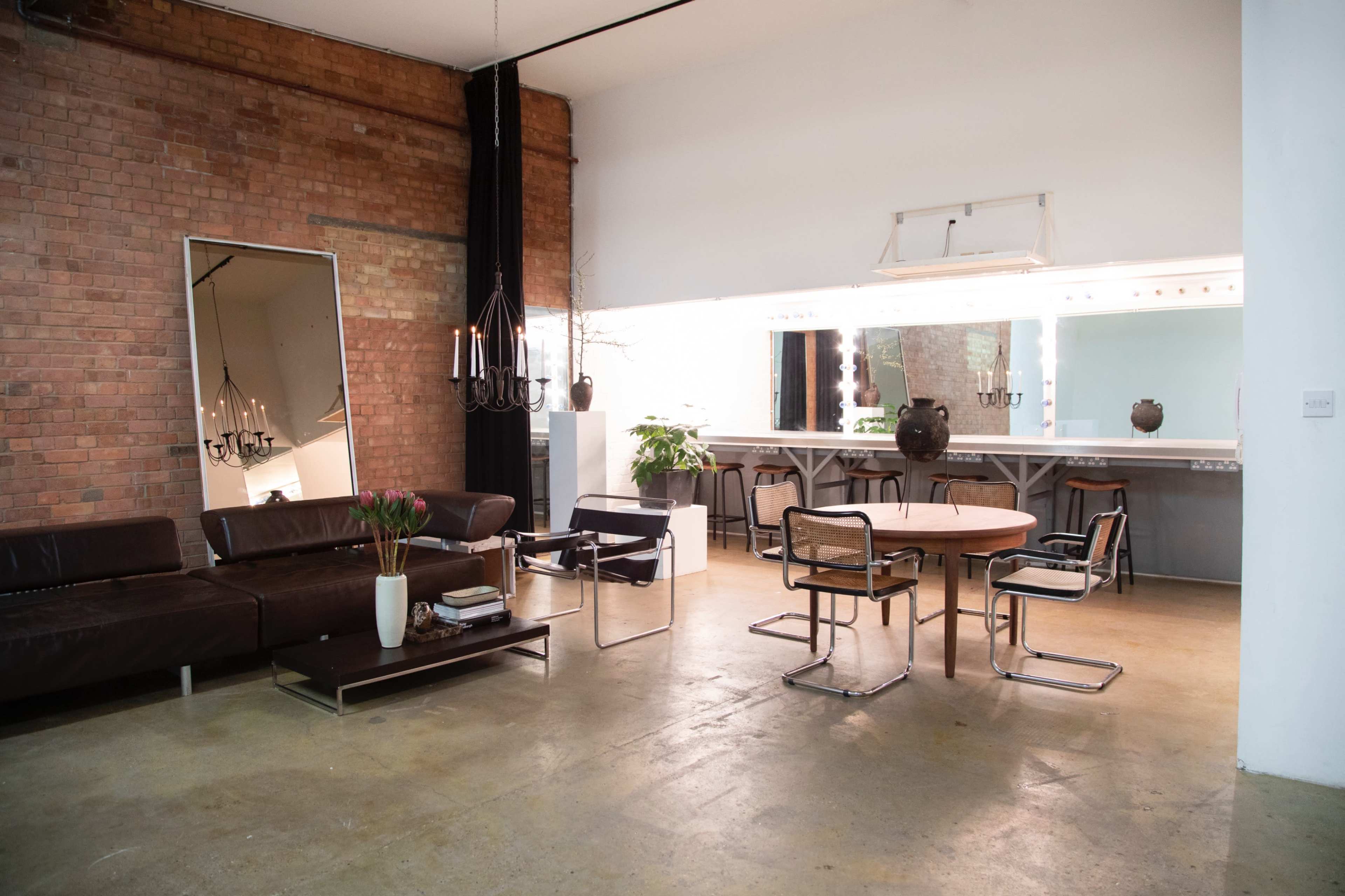 The scene features a spacious room with exposed brick walls, a brown leather sofa, a round wooden table surrounded by modern chairs, and a large mirror reflecting the minimalist decor.