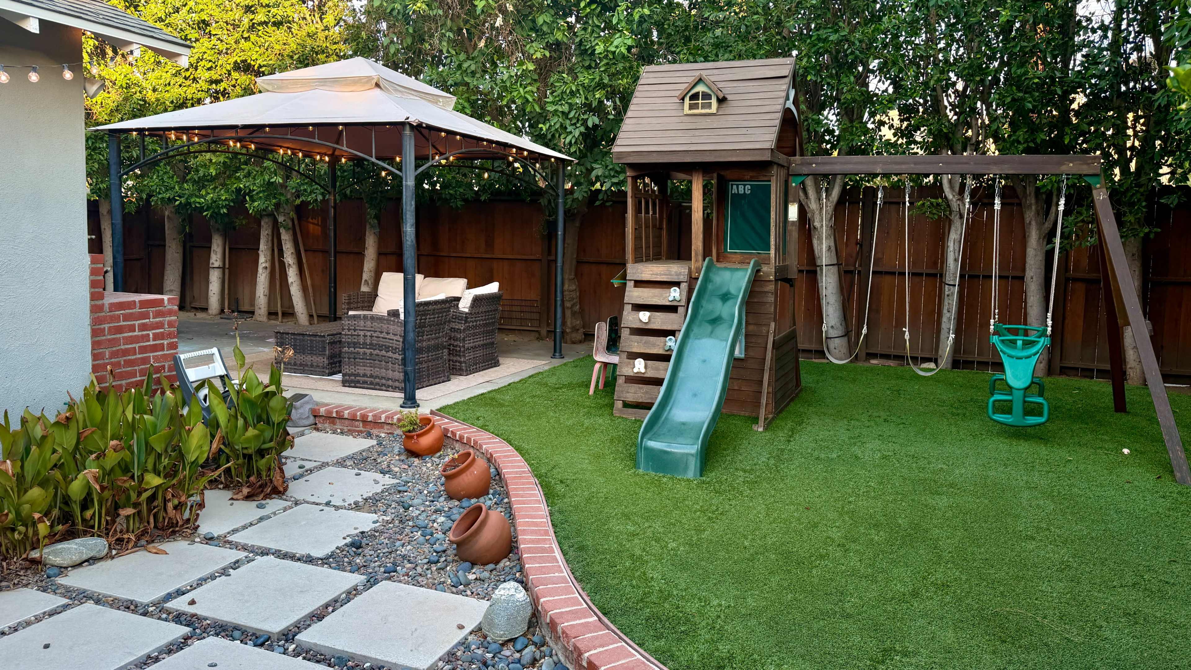 The image shows a backyard featuring a wooden playset with a slide and swings, a gazebo with seating, and a landscaped area with decorative pots and greenery.