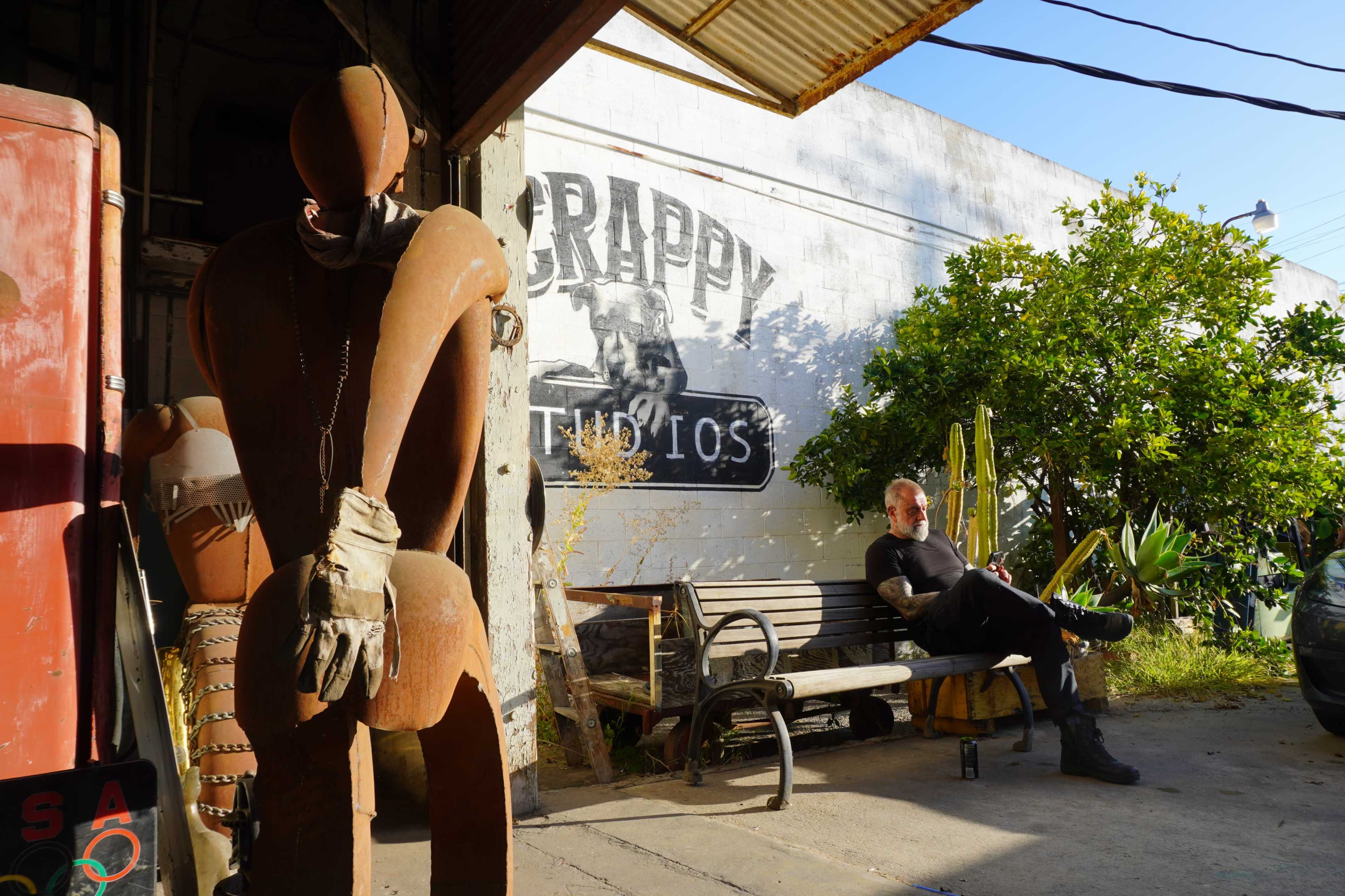 A rusted mannequin stands to the left while a man sits on a bench beside a wall that reads "CRAPPY STUDIOS" in a sunlit urban setting.
