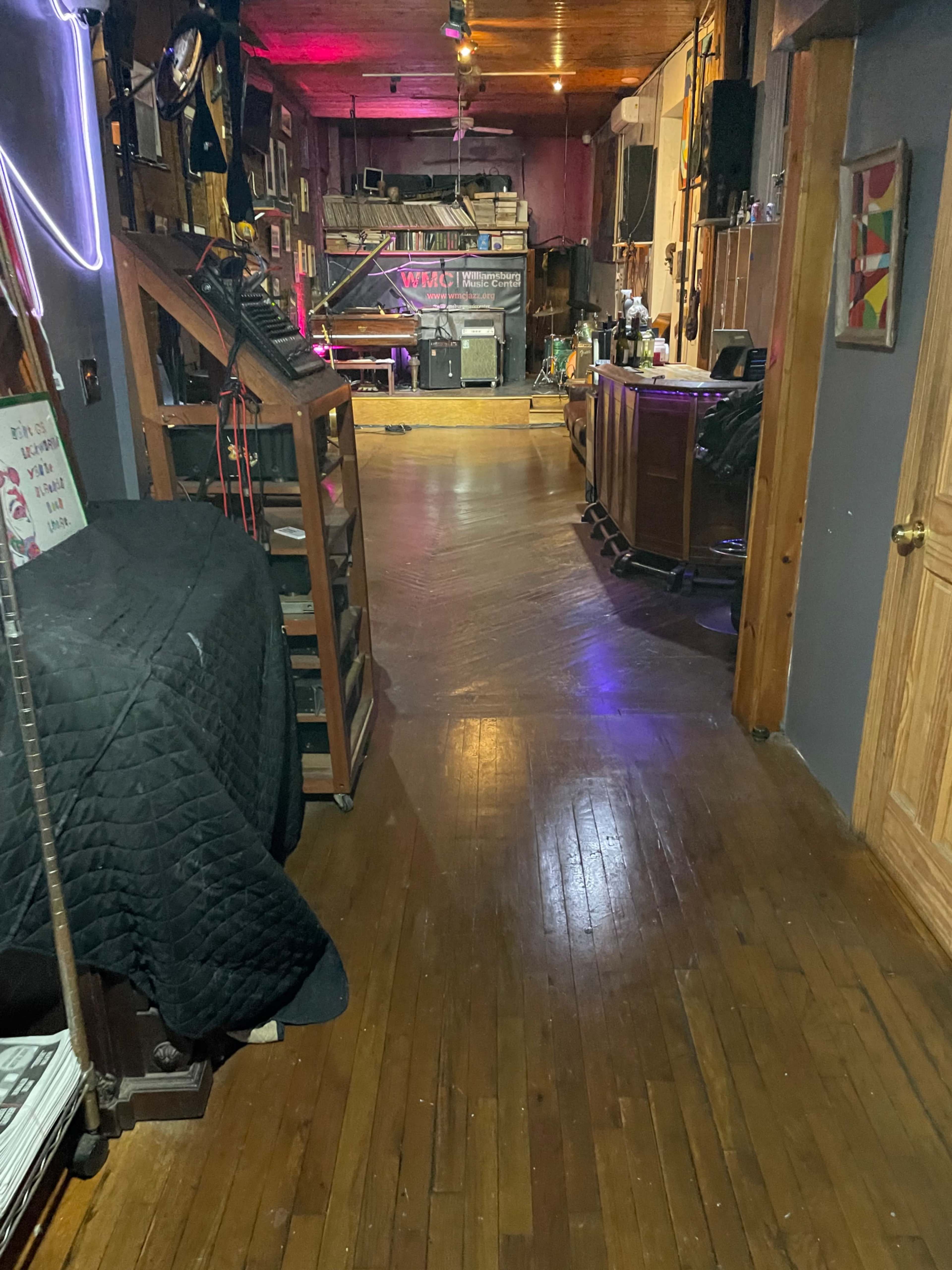 The image shows a long, narrow hallway leading to a stage area with wooden floors and various equipment and furnishings along the sides.