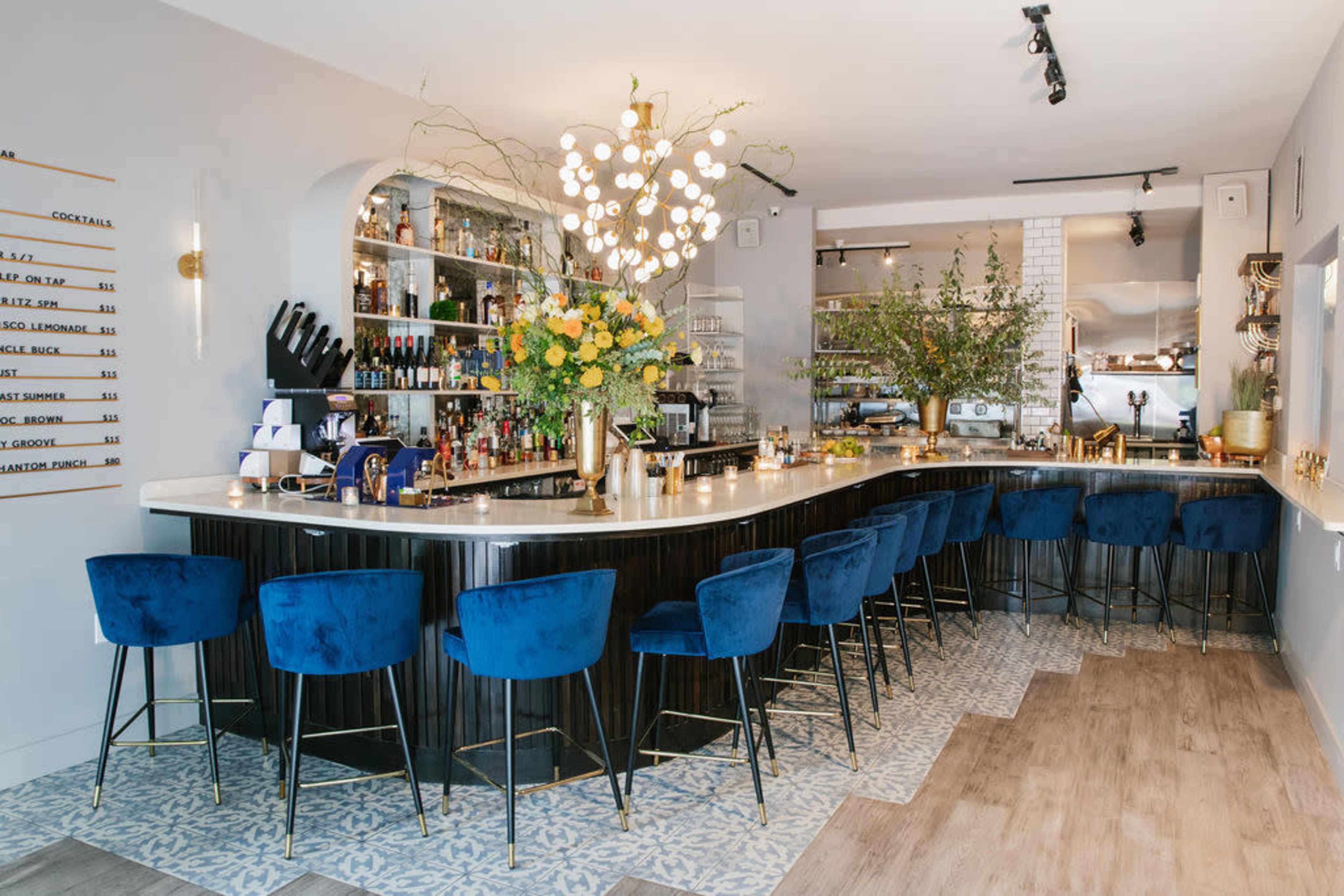 The image depicts a stylish bar area featuring a curved counter with blue upholstered stools, a well-stocked shelf of spirits, and decorative floral arrangements.