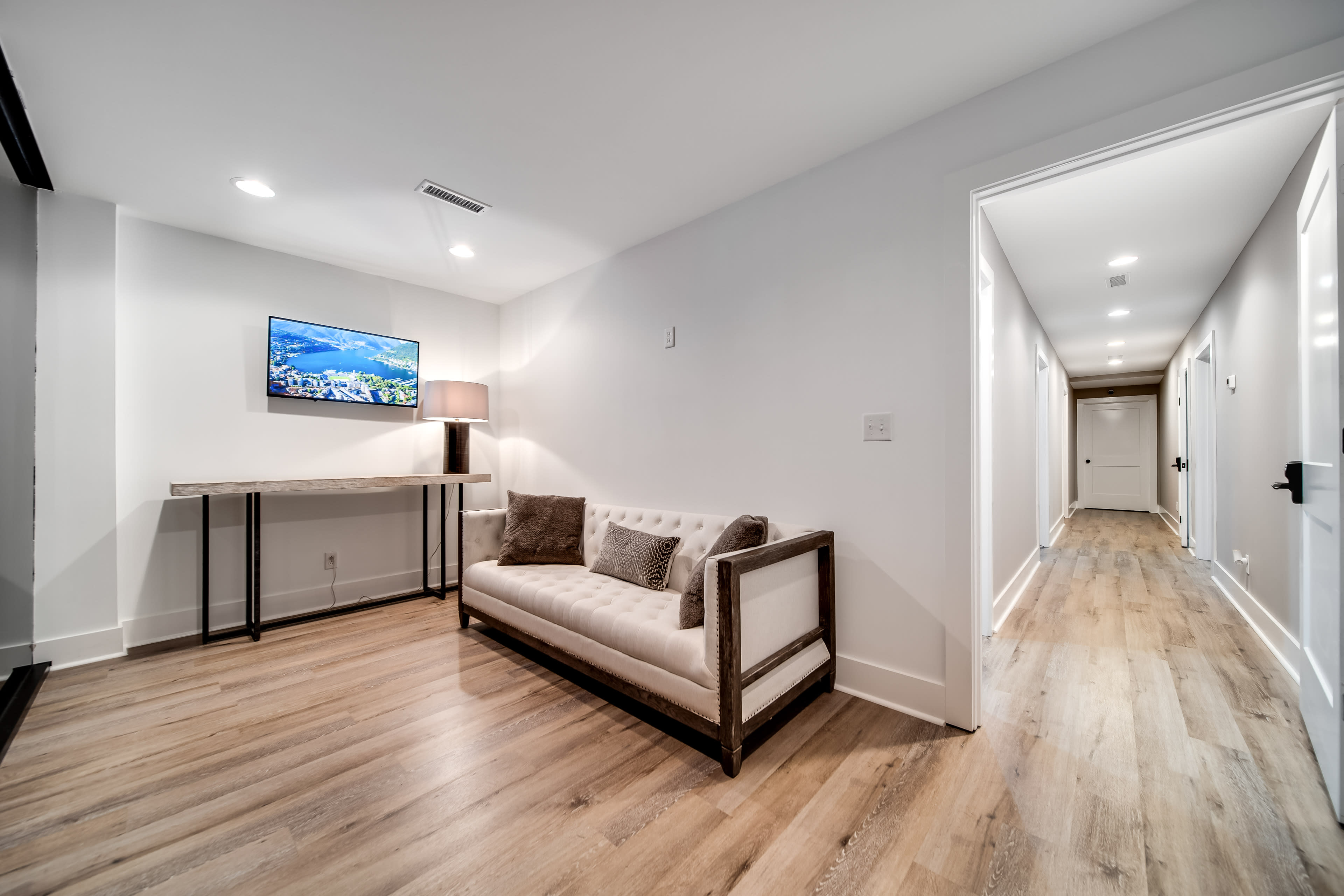 The image shows a narrow hallway with a modern couch, a TV mounted on the wall, and lit areas leading to a door at the end of the corridor.