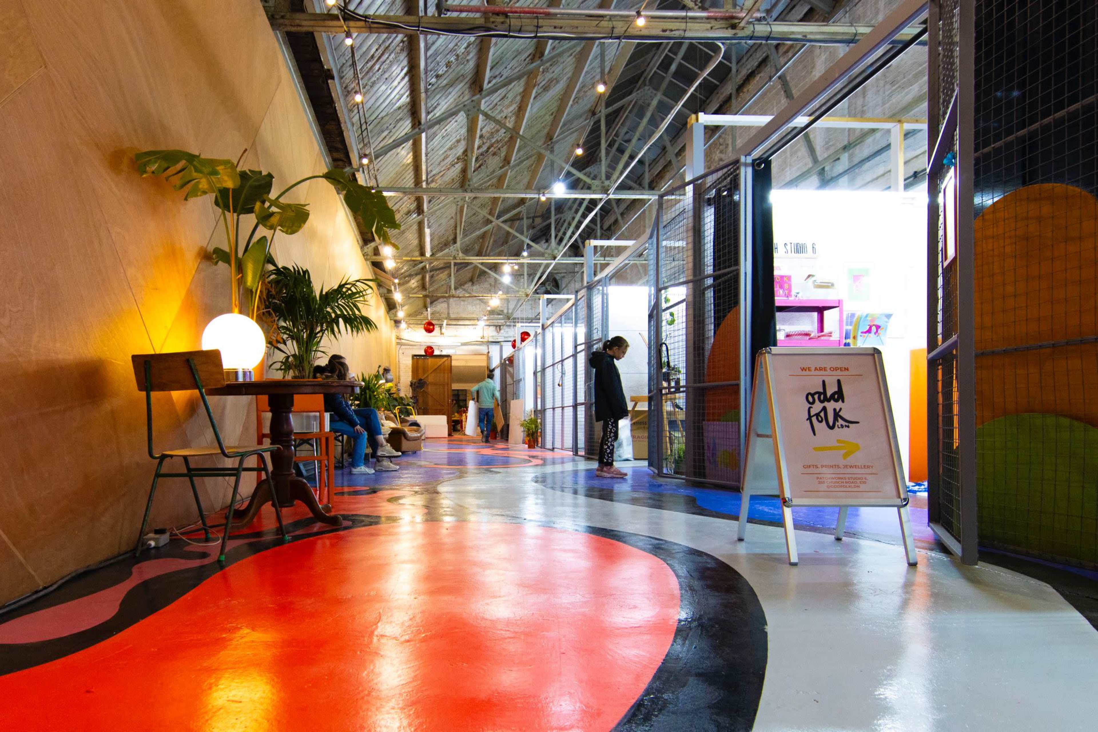 The image shows a colorful hallway with a mix of painted floors and plants, featuring tables and signs in a modern indoor space.
