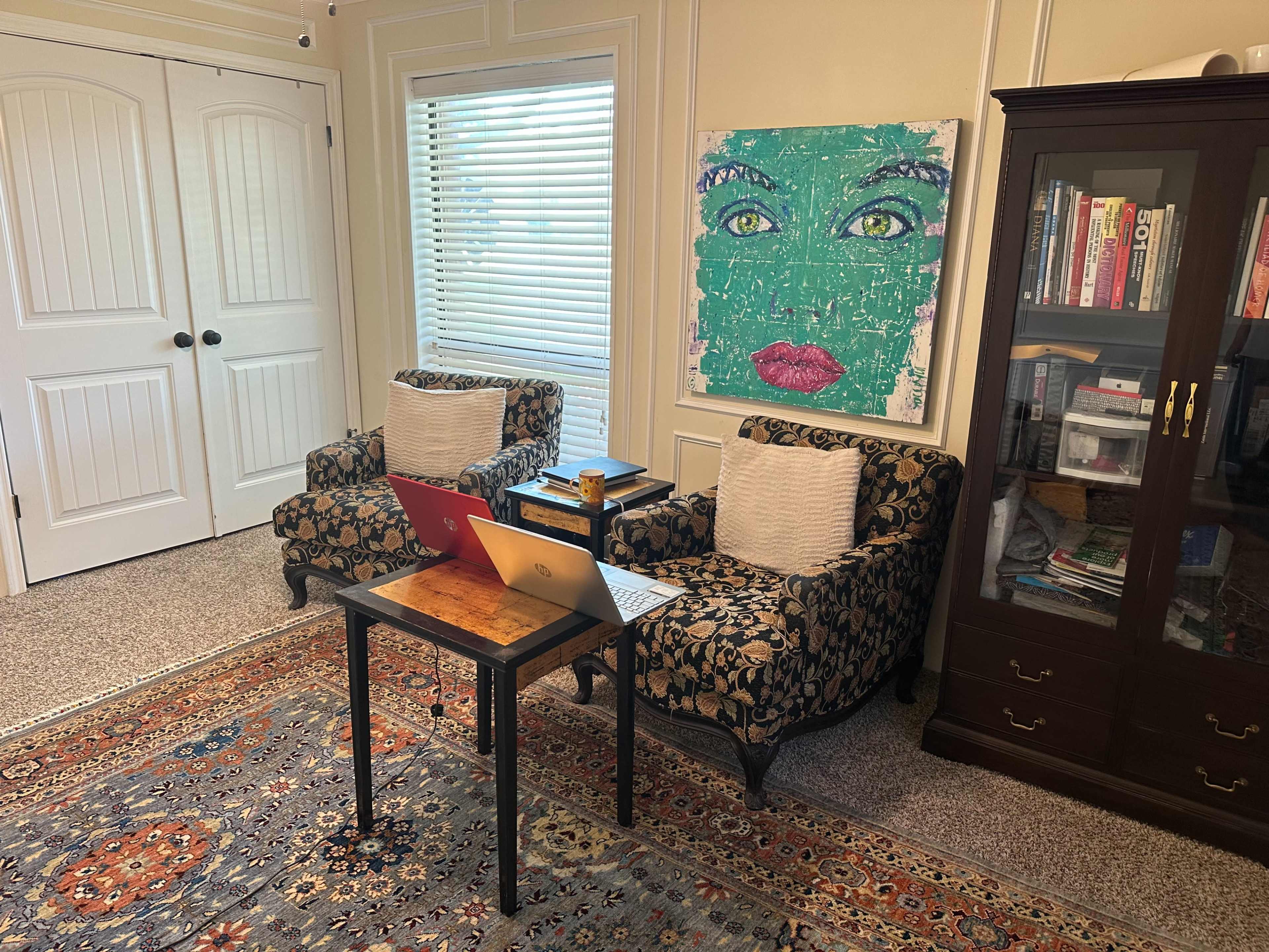 The image shows a cozy room with two patterned armchairs, a small table with two laptops, and a cabinet filled with books, all set against a backdrop of a colorful artwork on the wall.