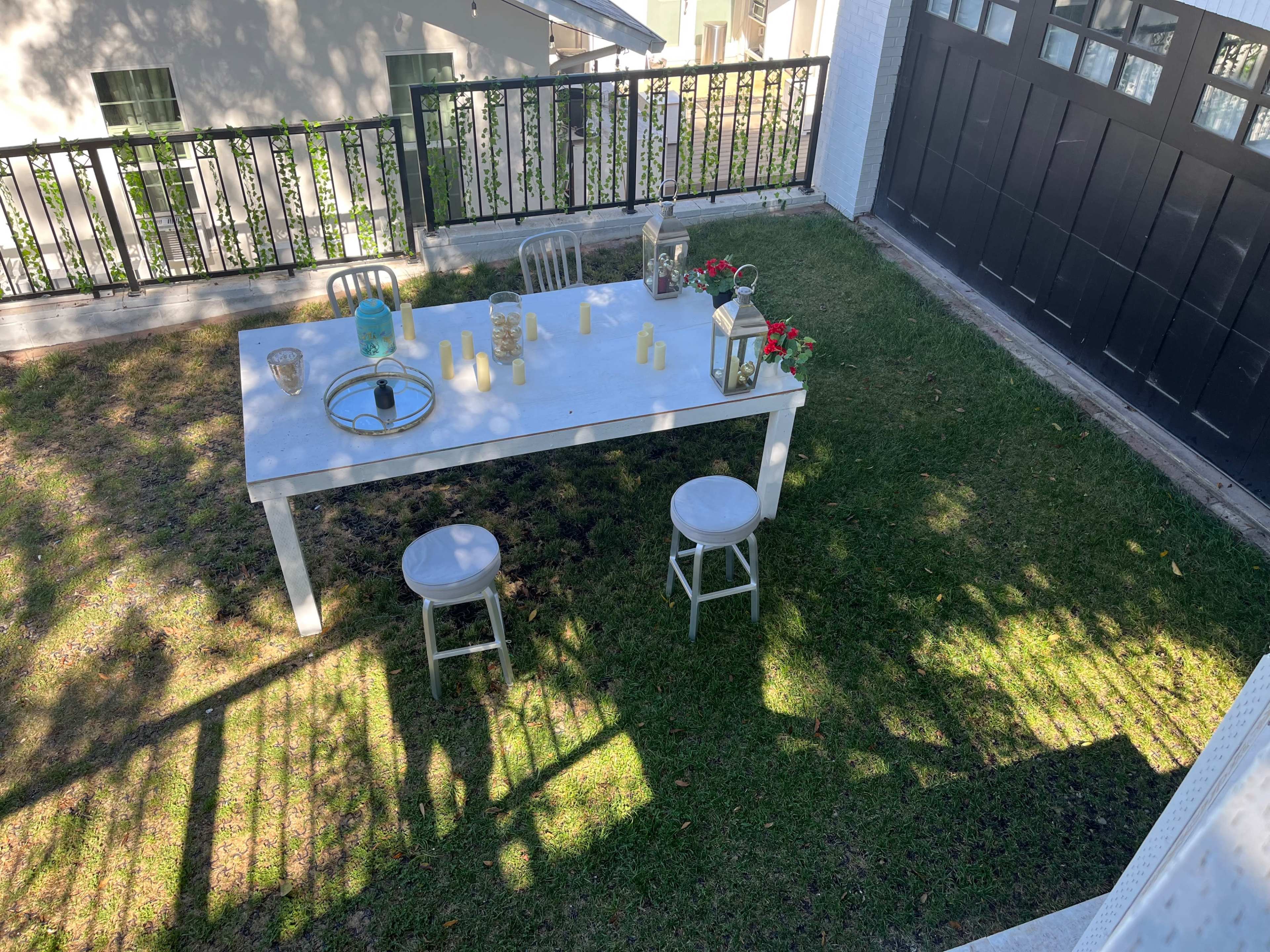 An outdoor dining table is set with candles and decor, surrounded by green grass and shadows cast from a nearby fence.