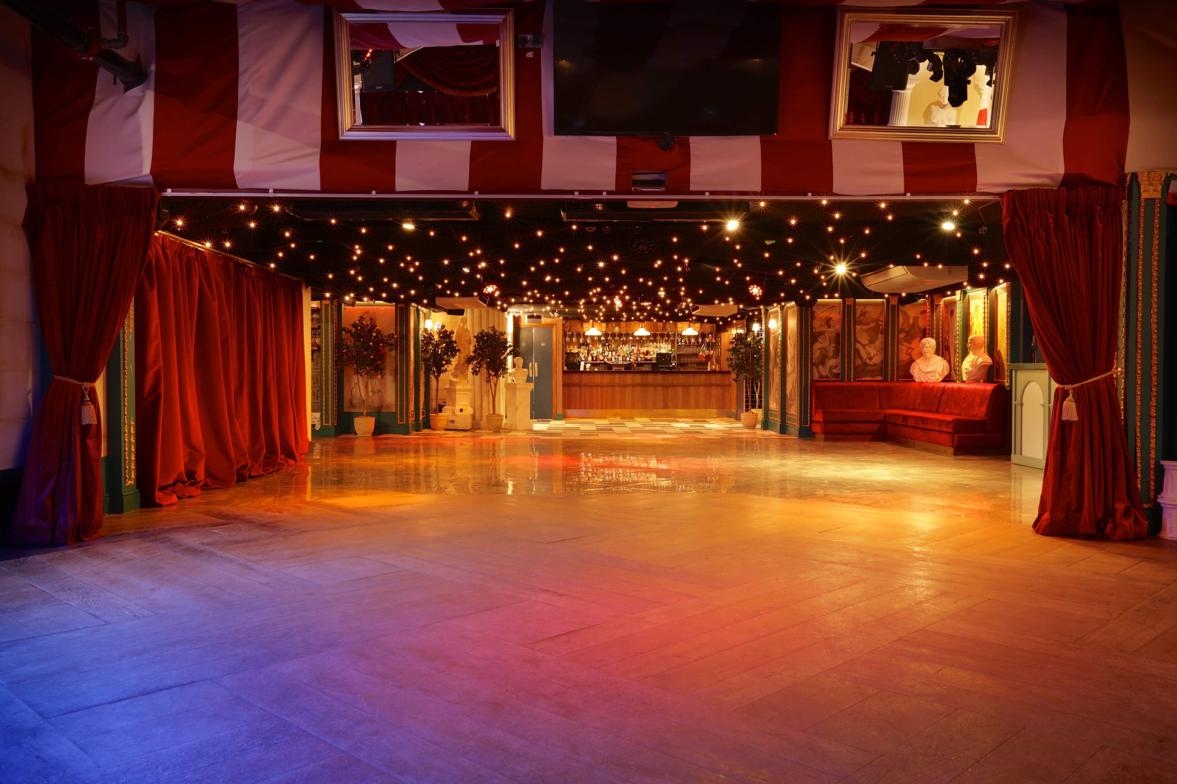 A spacious indoor venue featuring a polished wooden floor, red curtains, and soft lighting, with a bar area visible in the background.