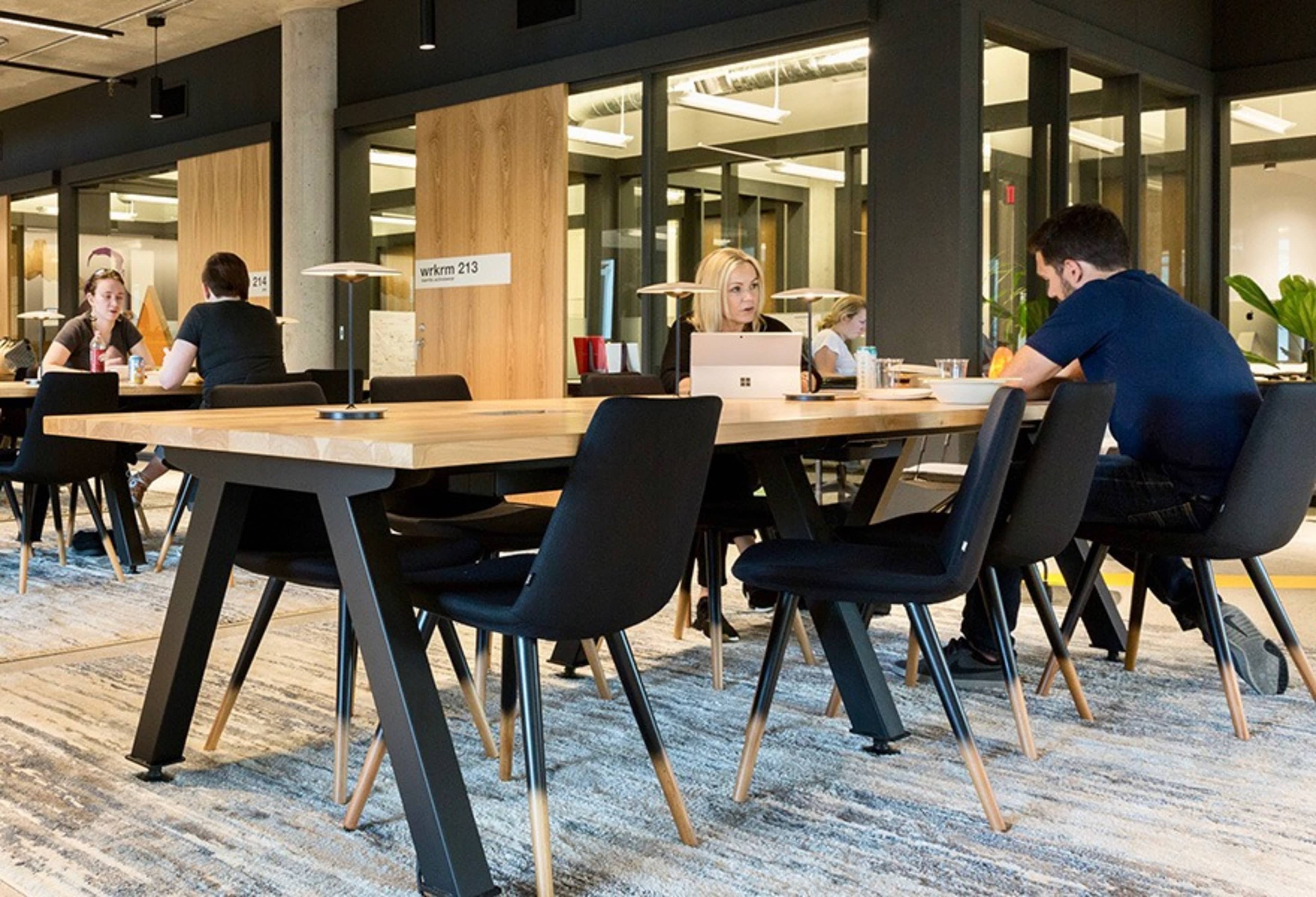 A modern office space features a large wooden table surrounded by black chairs, with several people engaged in work and discussions.