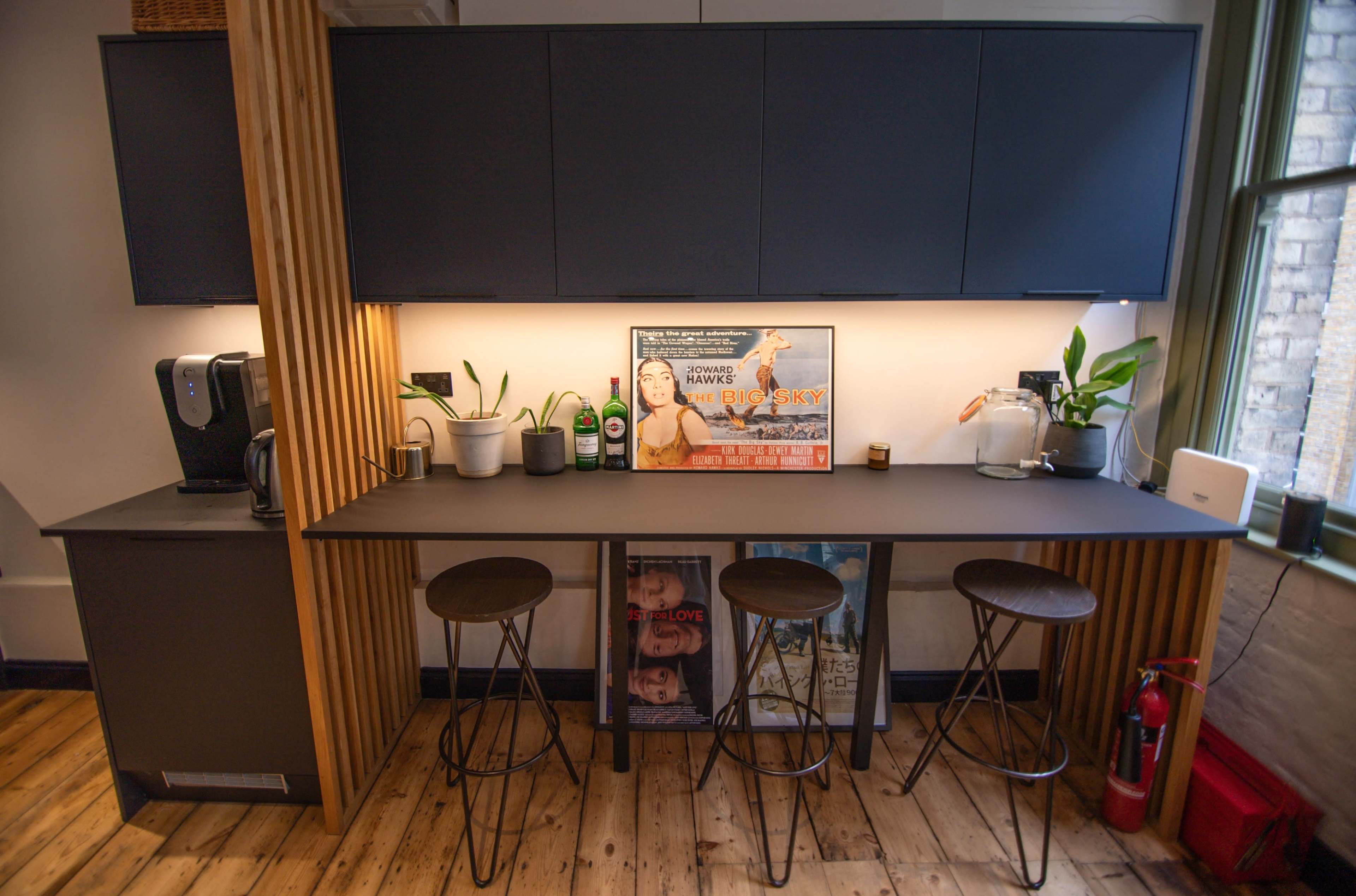 A modern kitchen with a long, dark countertop, three bar stools, and a coffee machine on the side, featuring a framed movie poster on the wall.