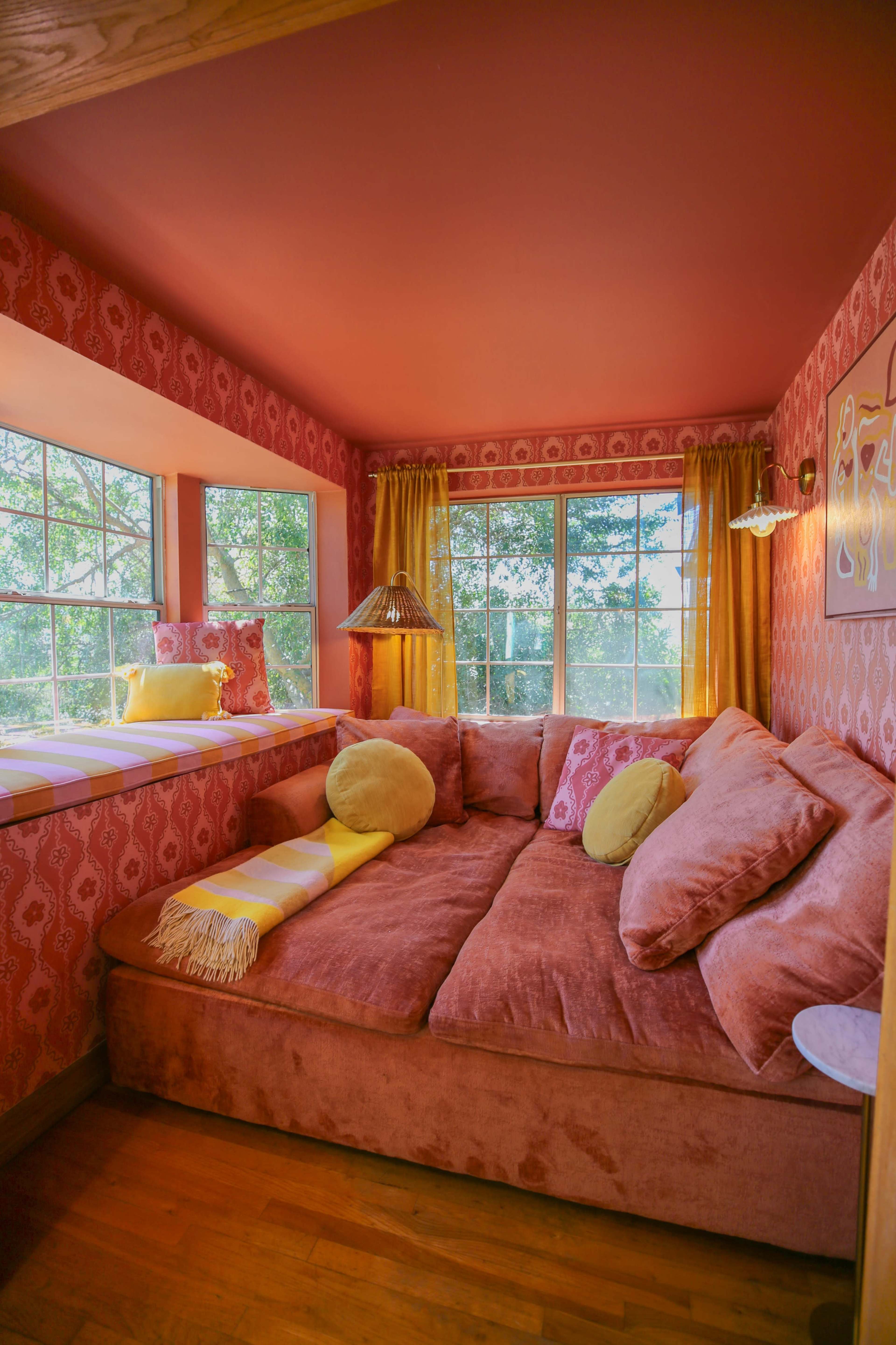 The image shows a cozy seating area with a large, plush couch surrounded by large windows that let in natural light, and walls adorned with patterned wallpaper.