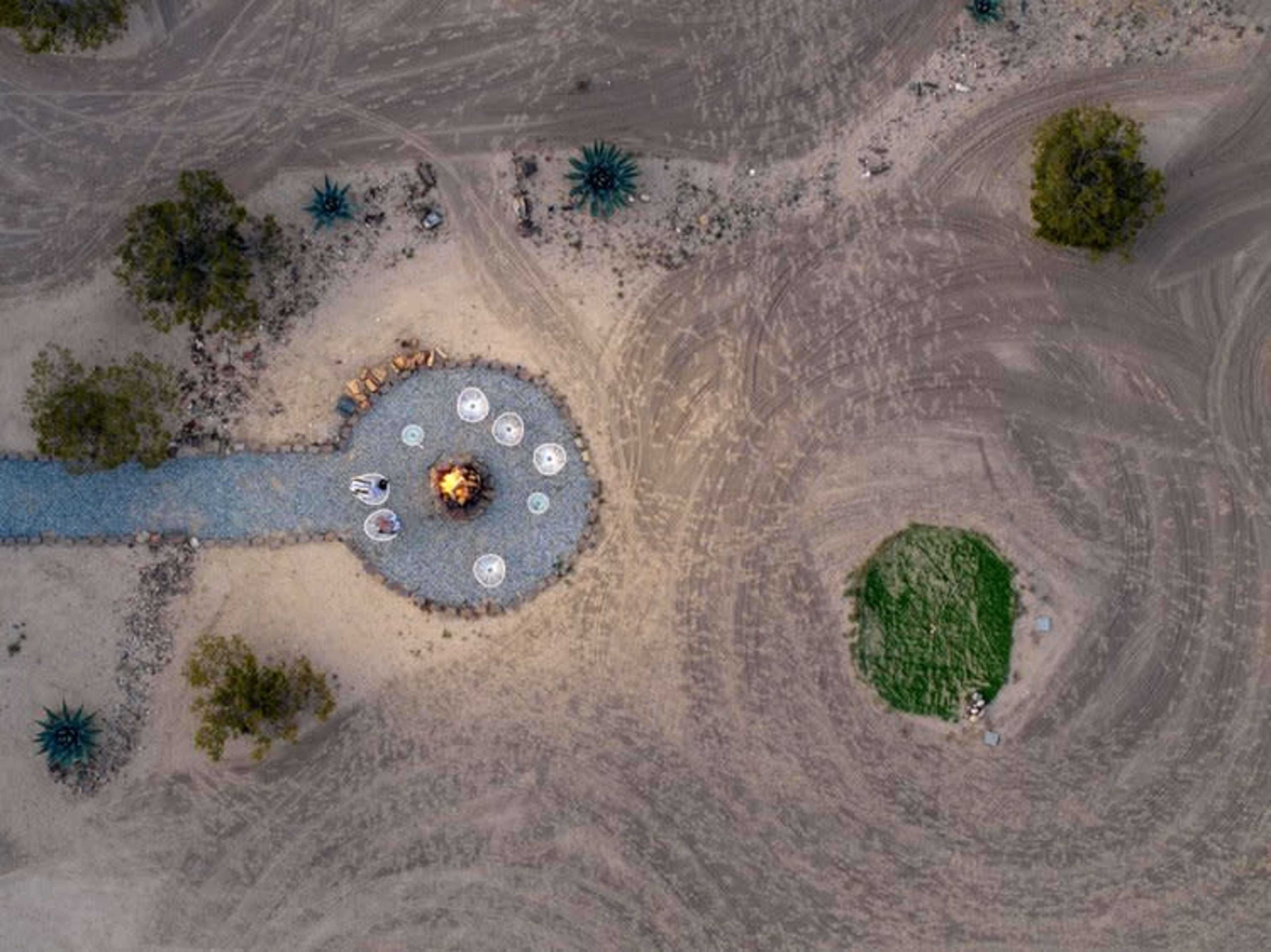 The image shows an aerial view of a circular gathering area with a fire pit surrounded by seating, bordered by a rocky path and desert terrain.