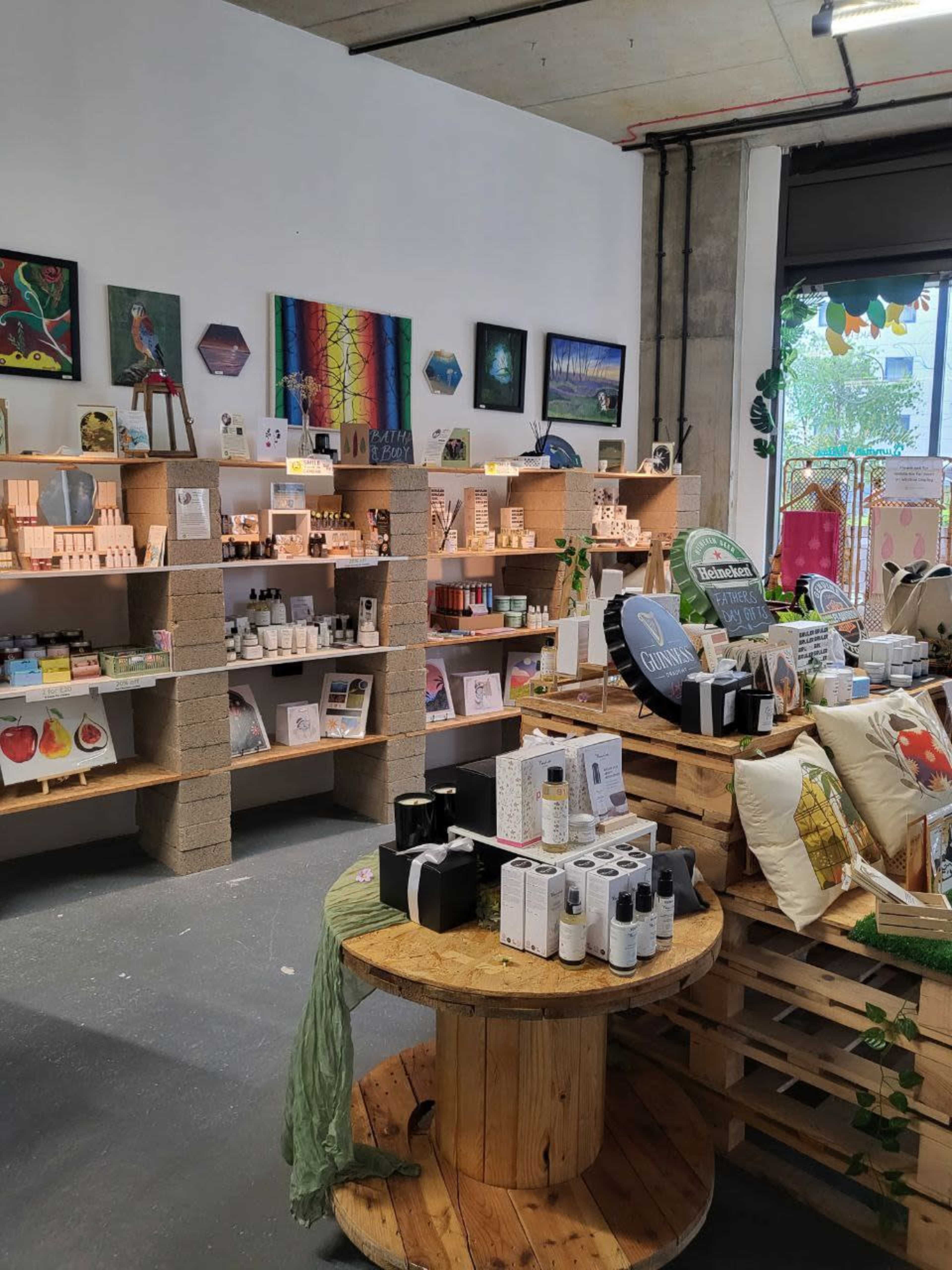 An indoor retail space with wooden shelves displaying a variety of products, including candles, cosmetics, and artwork, surrounded by plants and decor.