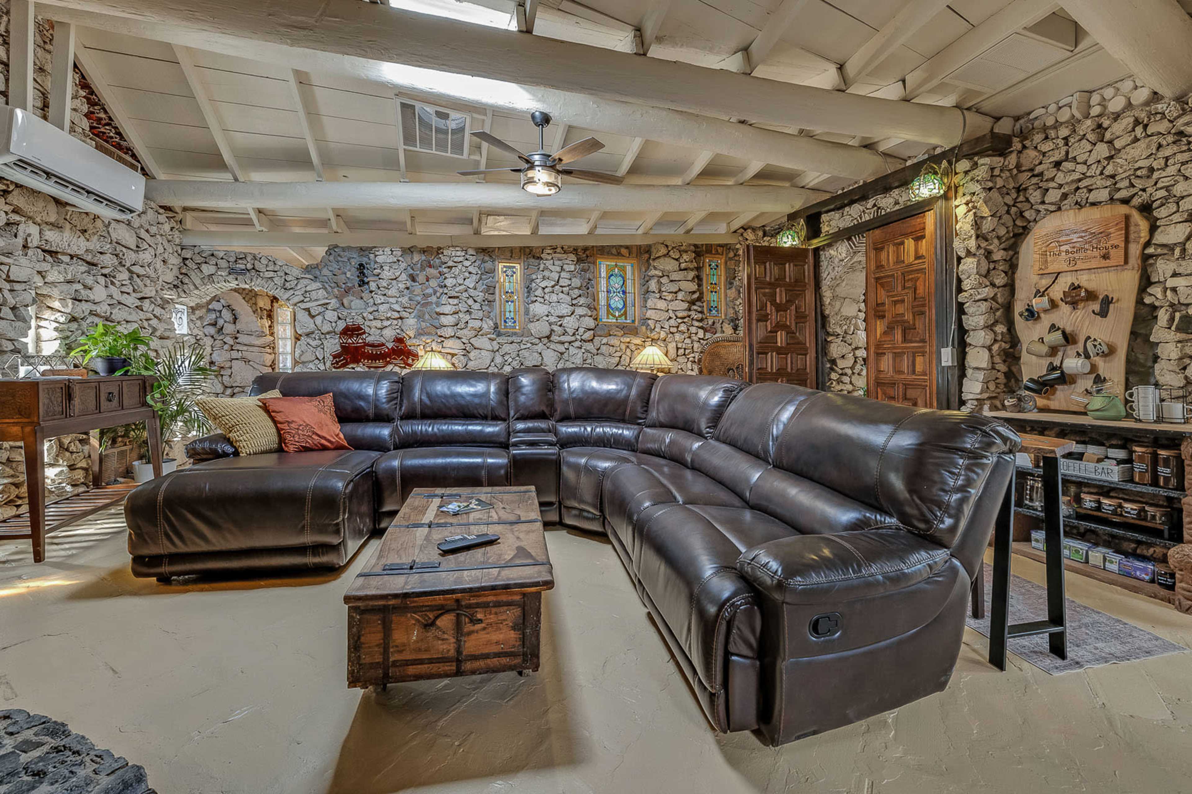 The image shows a spacious living room featuring a large brown leather sectional sofa, a wooden coffee table, and stone walls with decorative art and warm lighting.