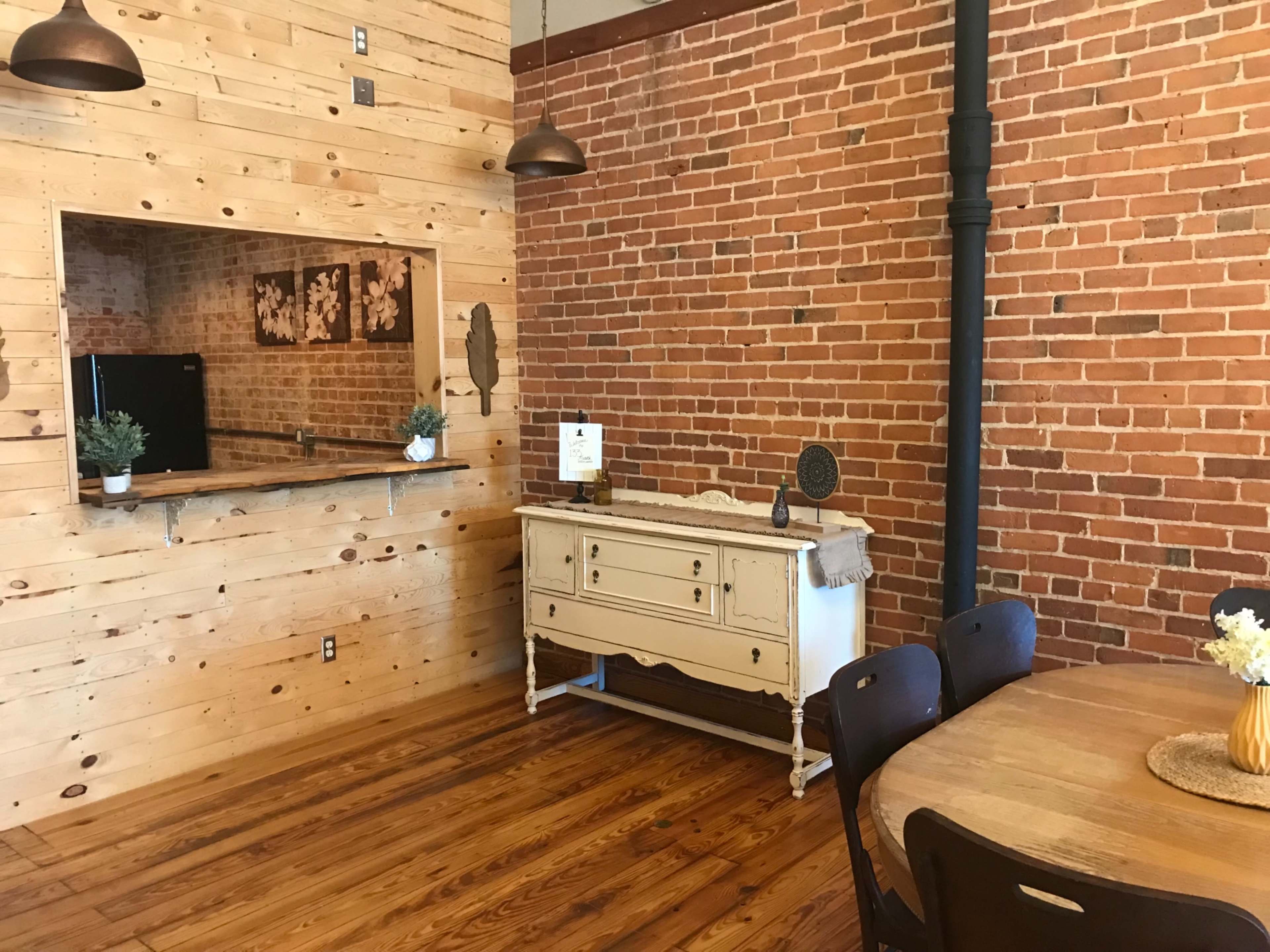 The image shows a dining area with a wooden table and chairs, adjacent to a kitchen area featuring a brick wall and a light-colored sideboard.