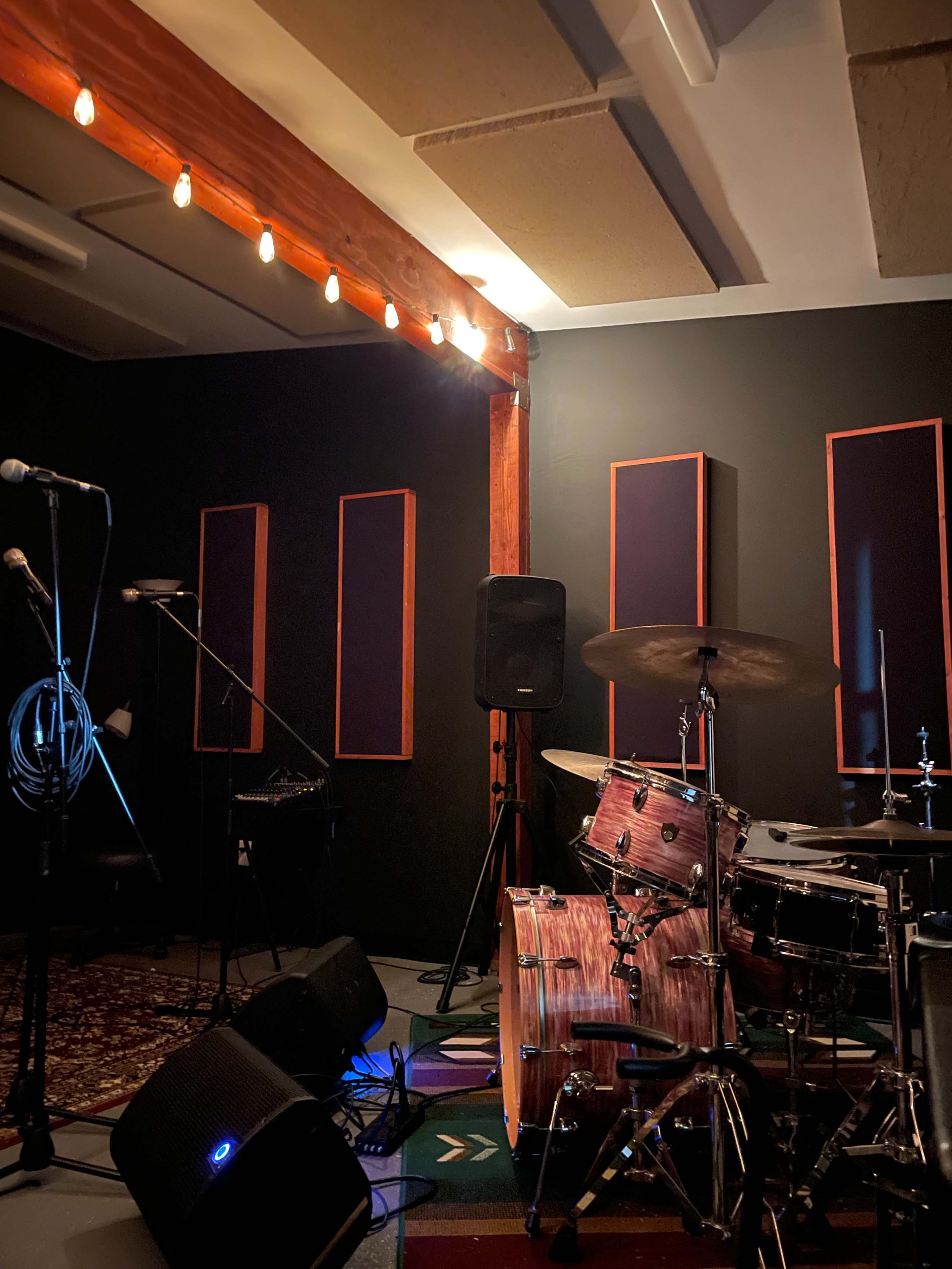 A music rehearsal space with a drum set, microphones, and sound equipment against black walls.