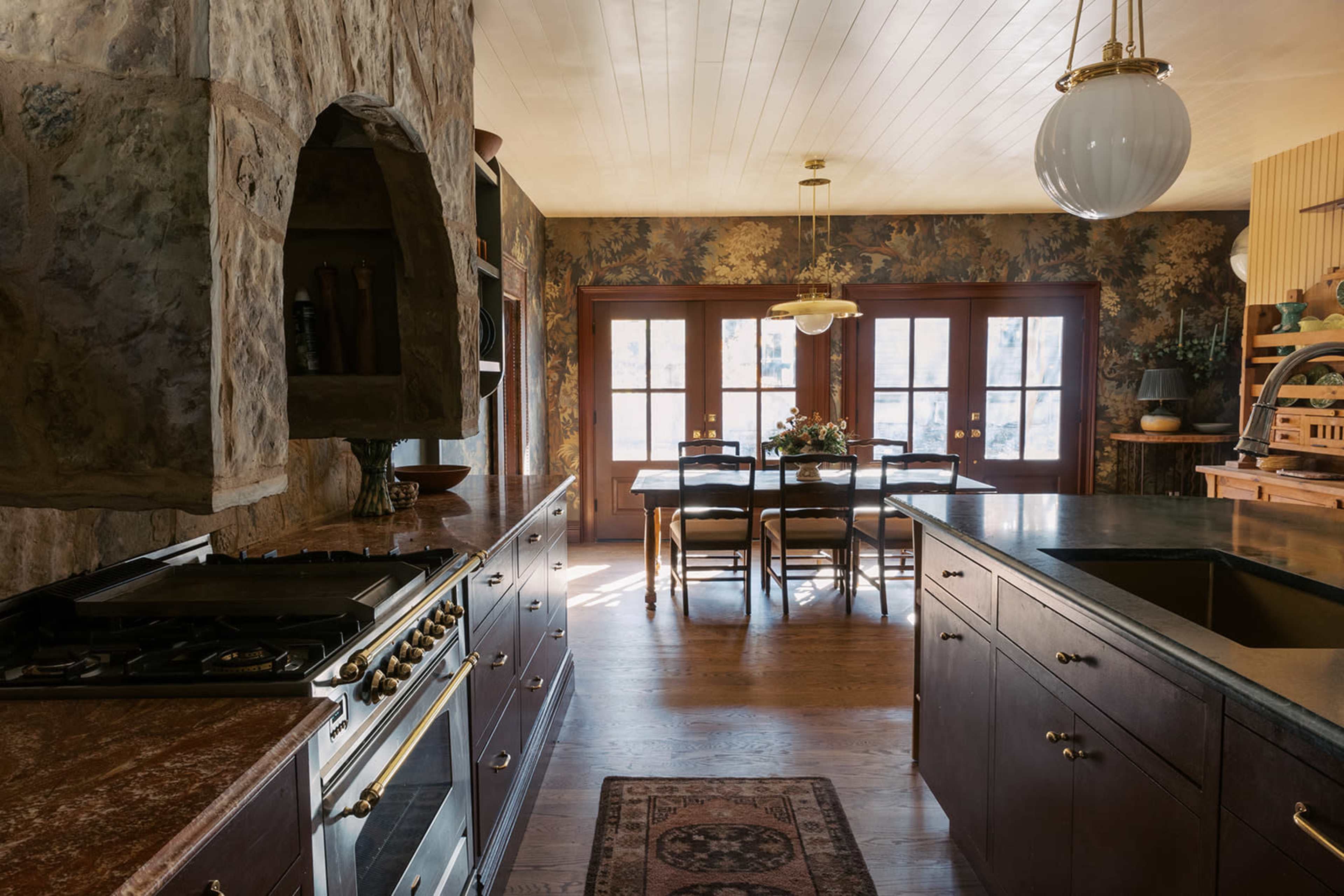 The image depicts a kitchen with stone walls and wooden cabinets, featuring a dining area with a table and chairs adjacent to large glass doors that open to an outdoor view.