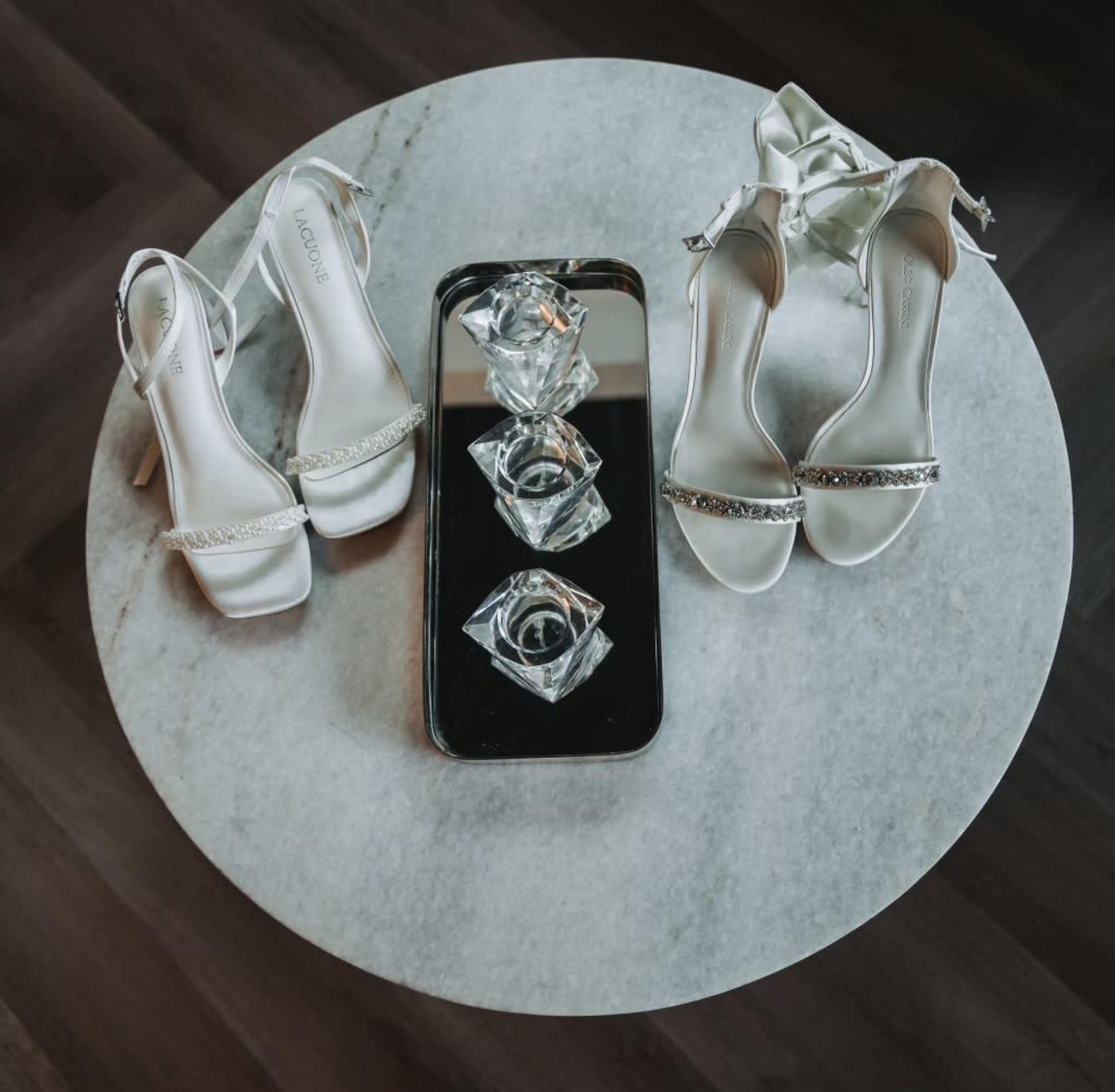 A pair of elegant white high-heeled shoes is placed on a round marble table beside a rectangular tray with three glass crystal decor pieces.