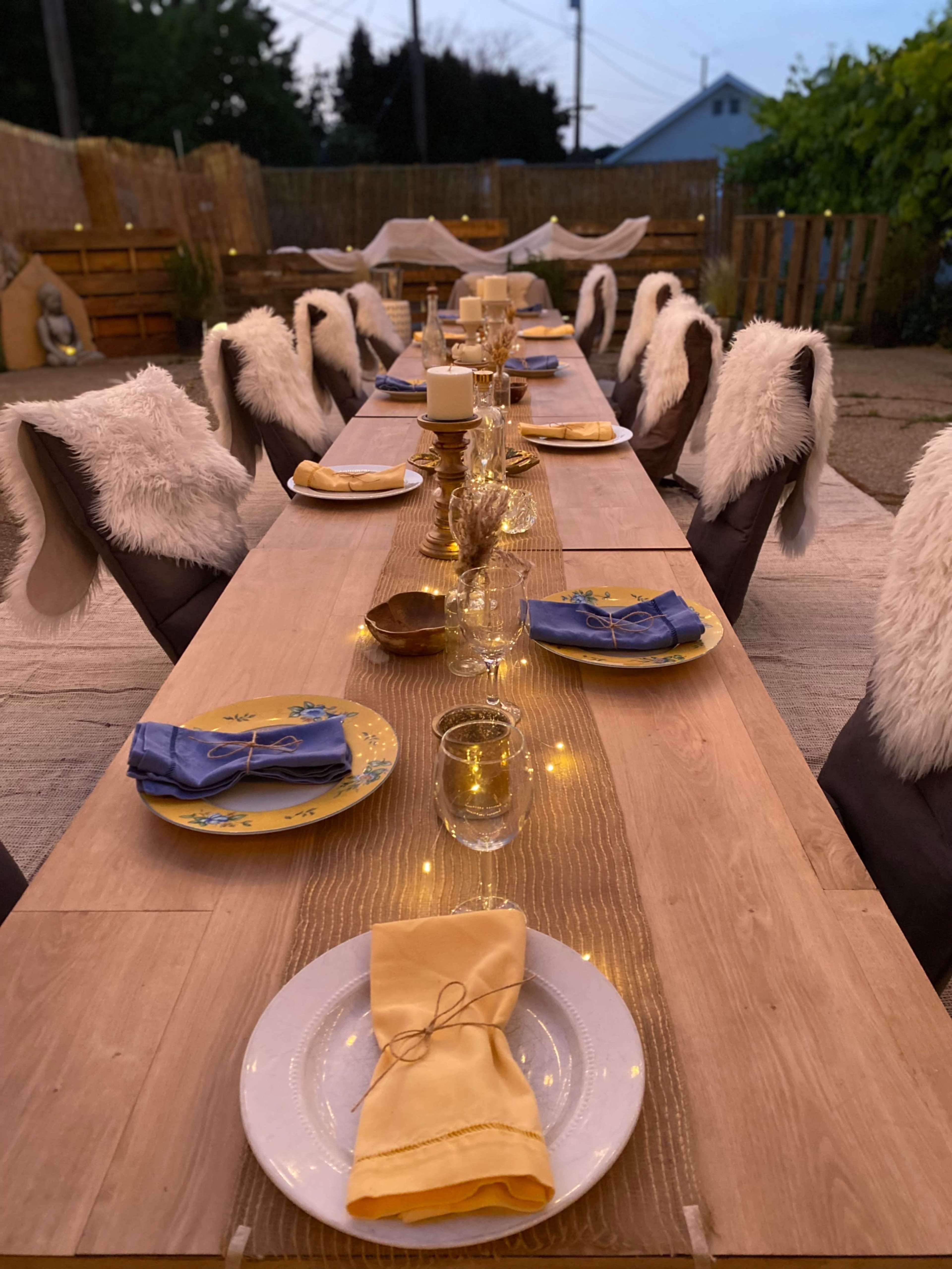 A long outdoor dining table is elegantly set with plates, glasses, and folded napkins, surrounded by chairs adorned with white fur throws.