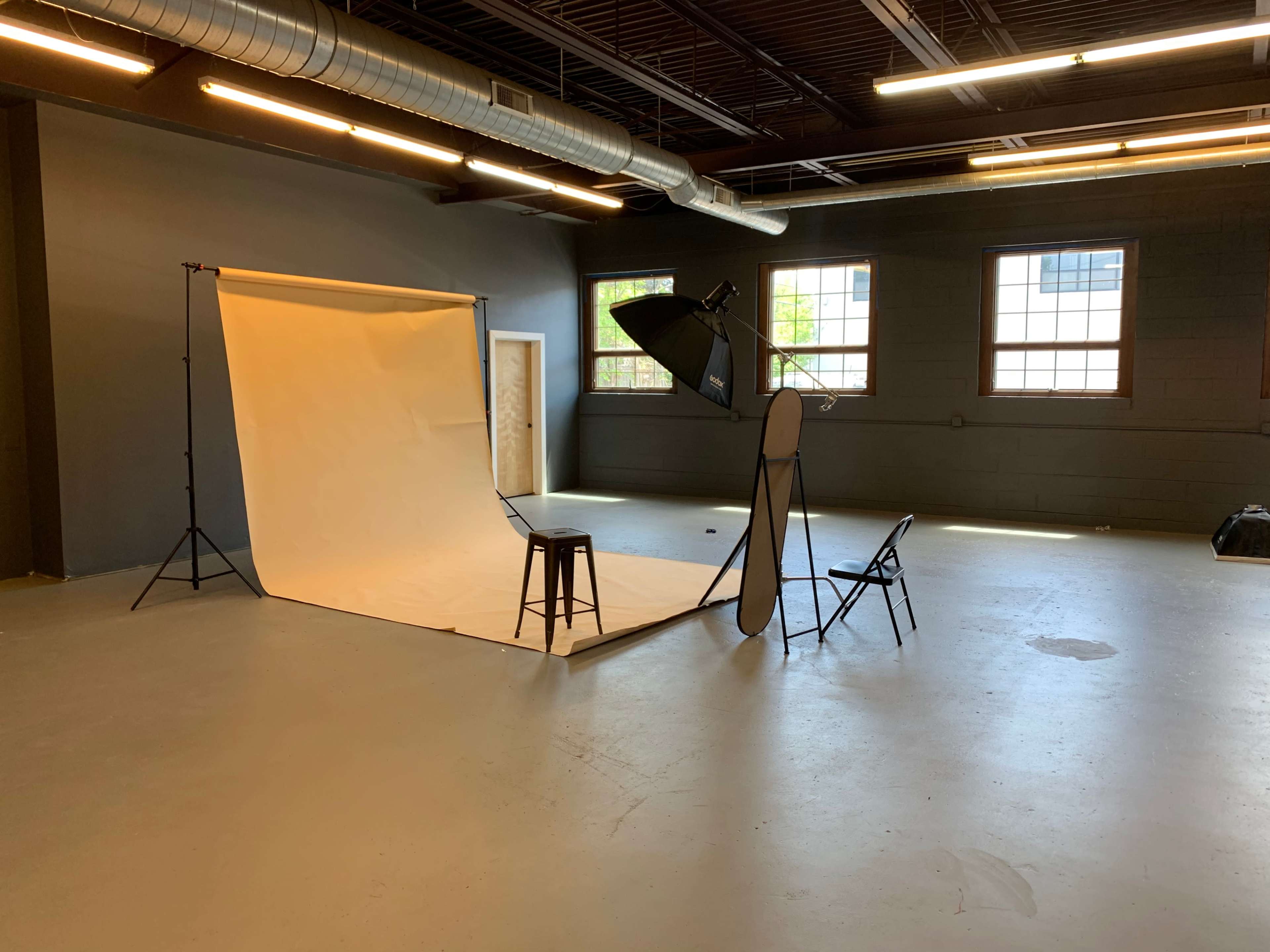 A photography studio setup with a backdrop, lighting equipment, a stool, and chairs arranged on a concrete floor.