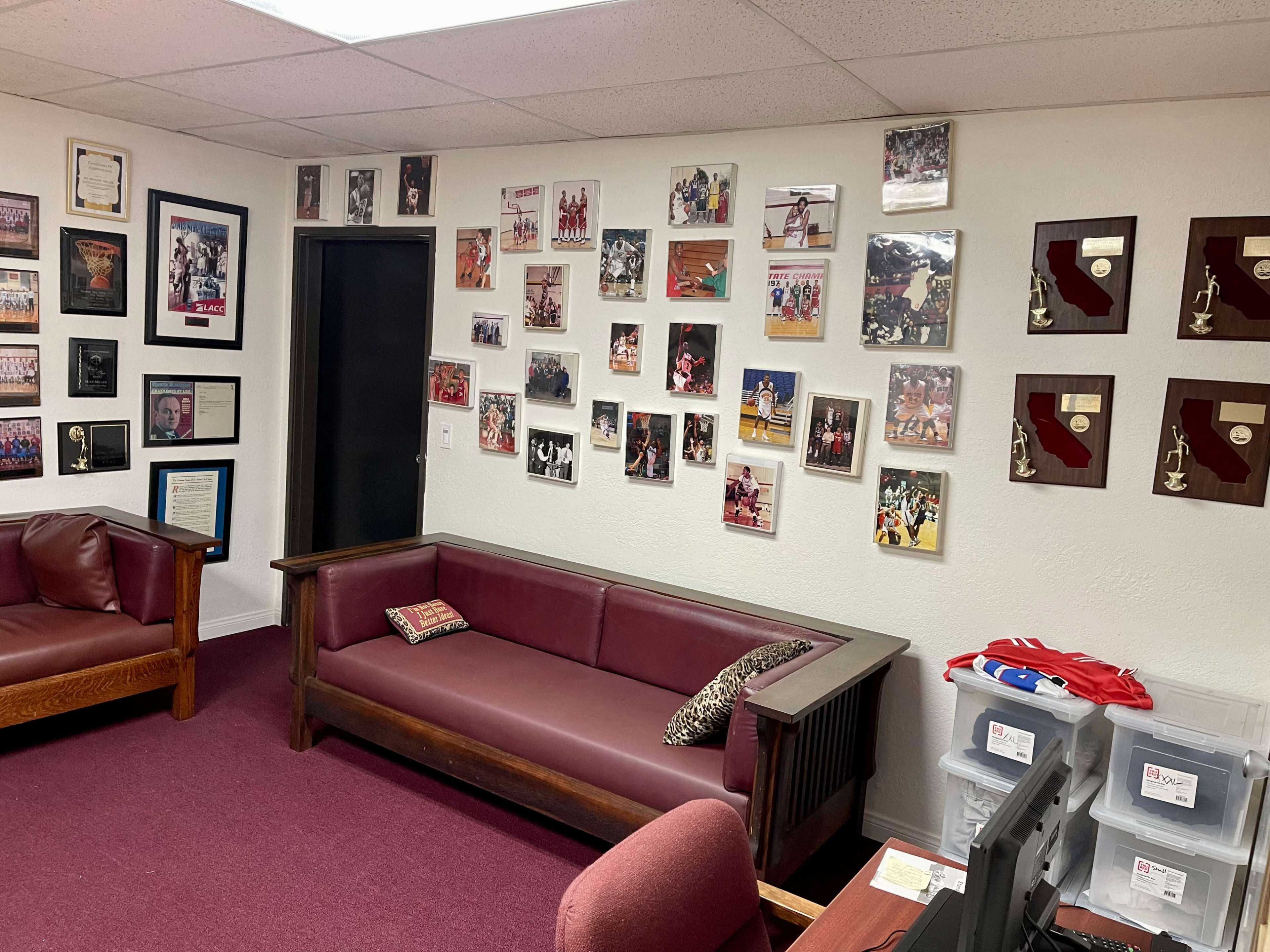An office space with a maroon carpet, two wooden couches, a desk with a chair, and walls decorated with framed photos and awards related to sports.