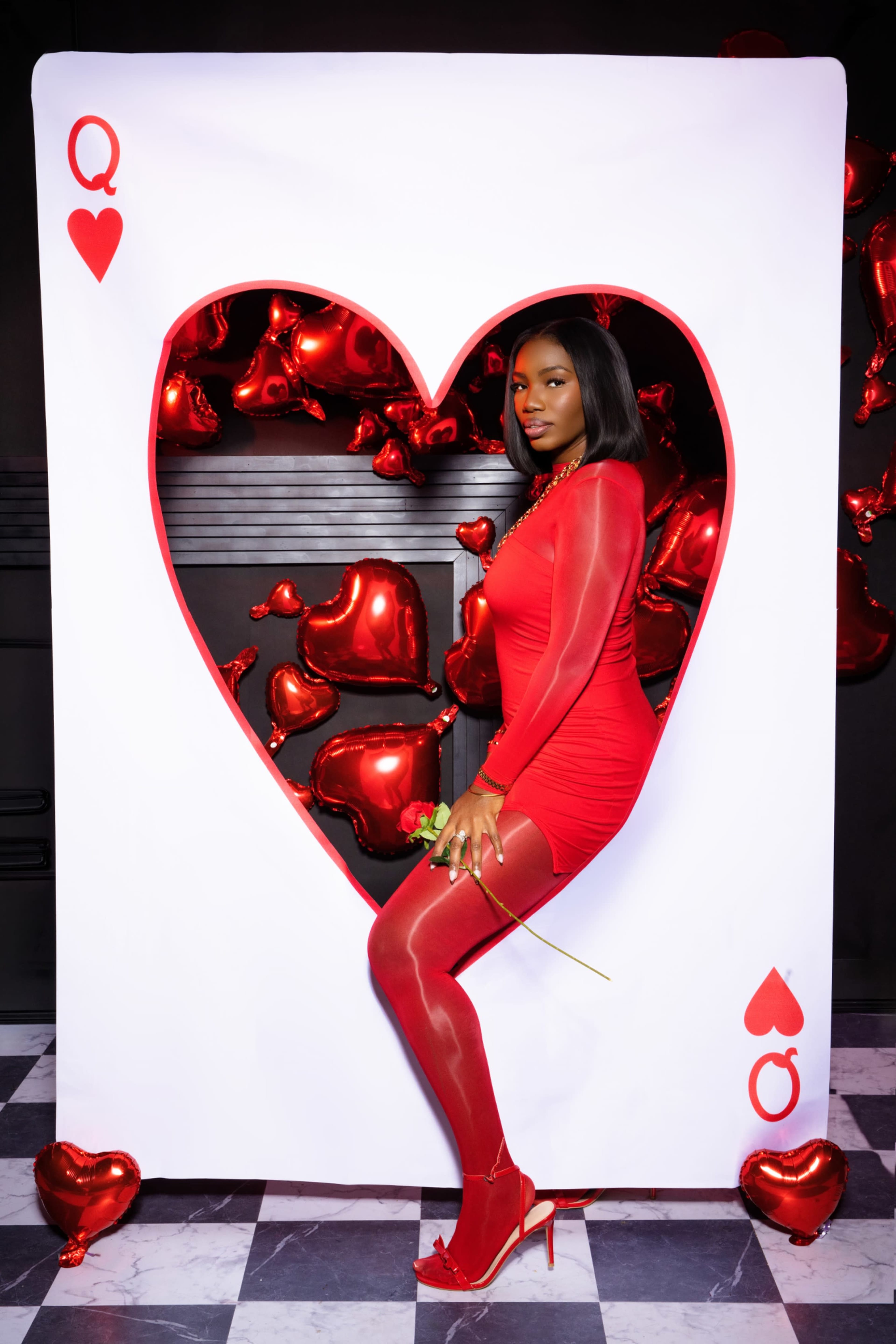 A woman in a red dress poses inside a large heart-shaped cutout decorated with red balloons.