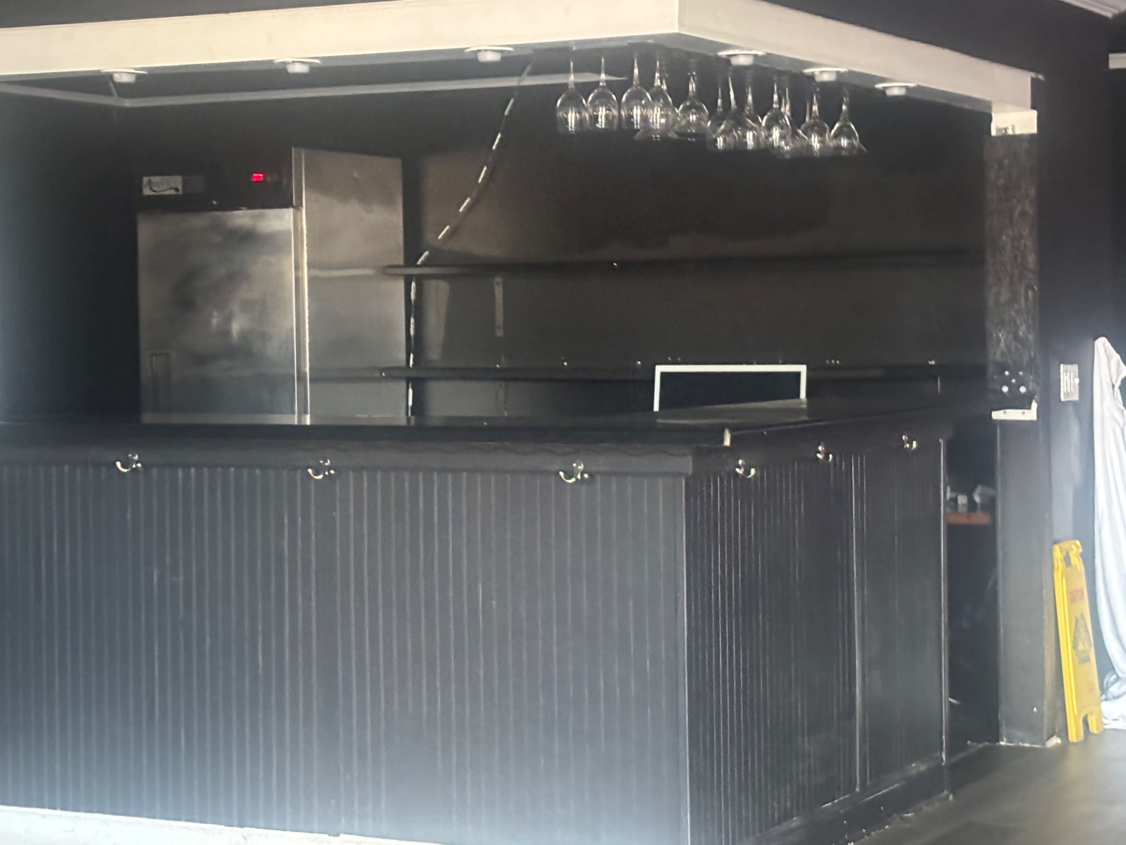 The image shows a darkly colored bar area with a stainless steel refrigerator, shelves, and hanging glasses.