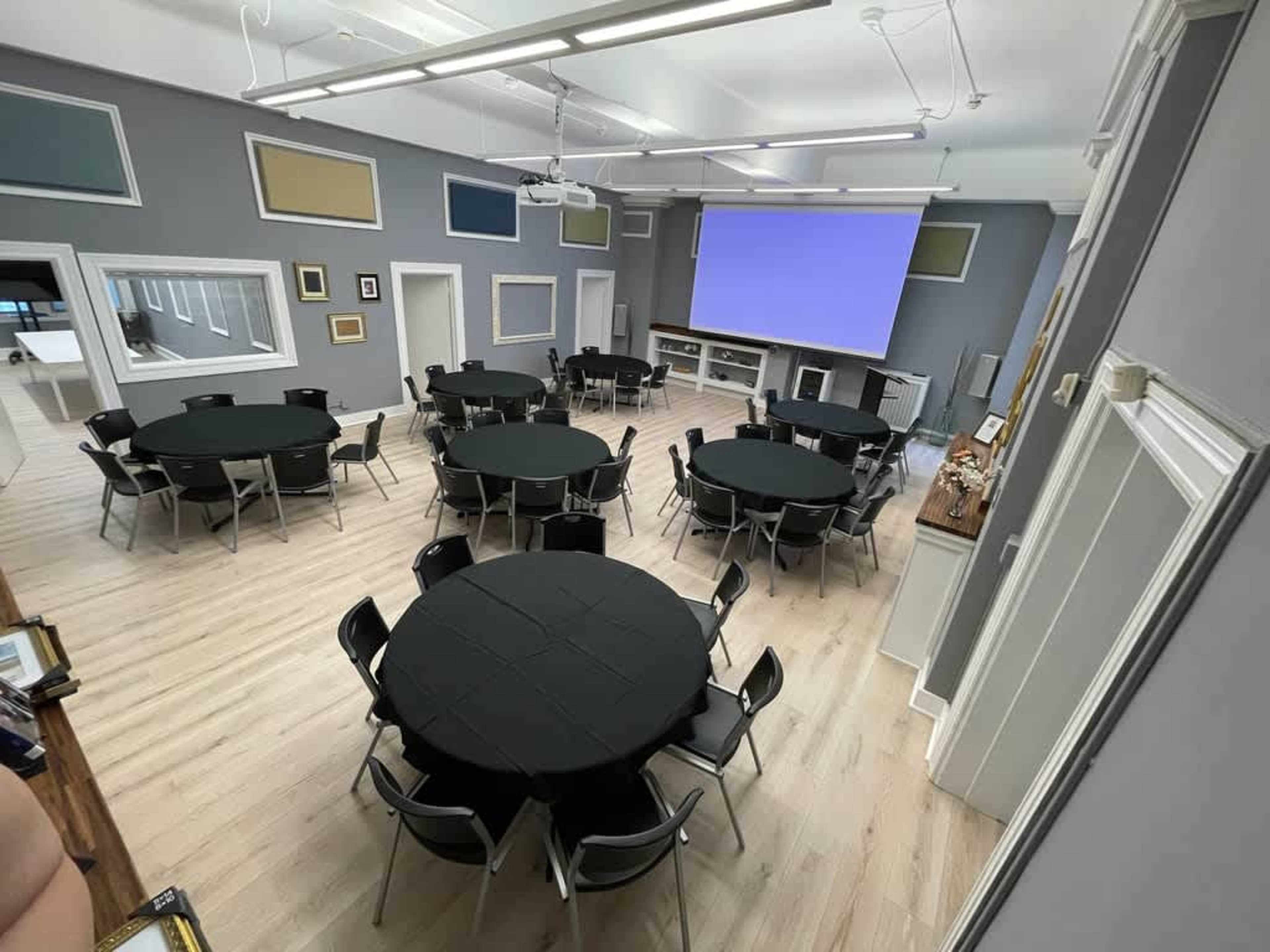 The image shows a large meeting room with several circular tables covered in black cloth, arranged around a projection screen at the front and gray walls adorned with colorful framed panels.