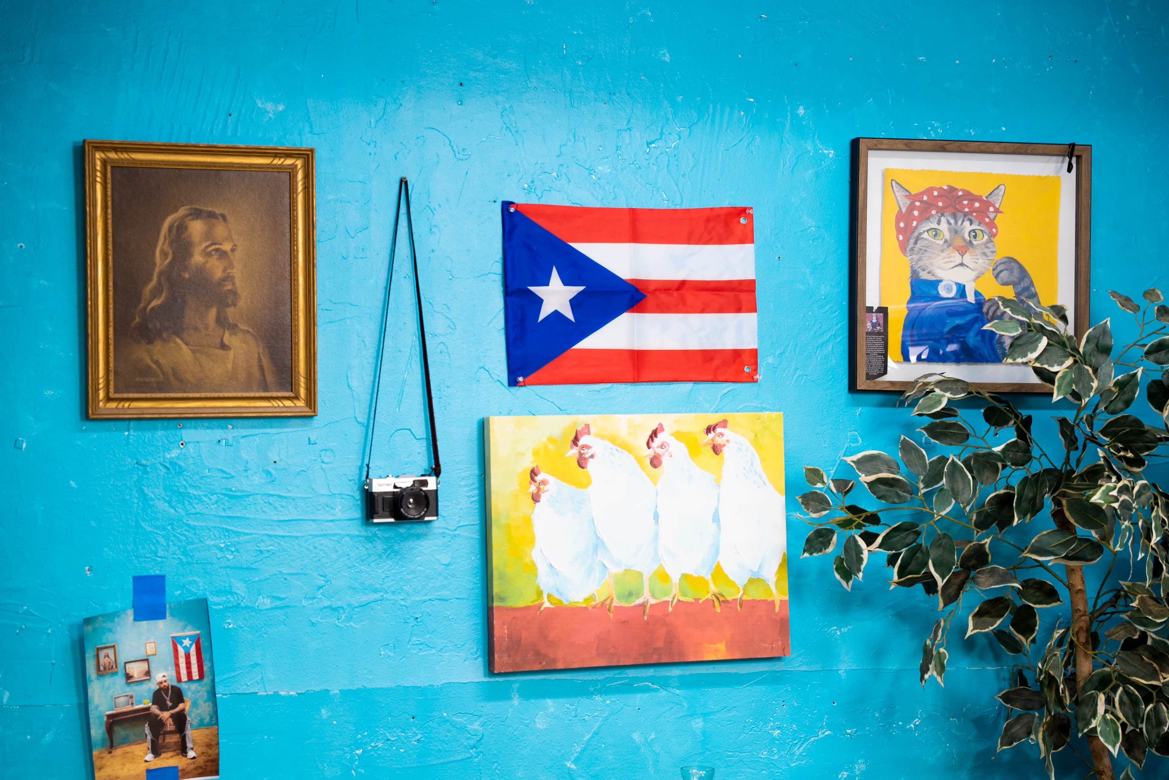 The scene features a turquoise wall adorned with a portrait of Jesus, the Puerto Rican flag, a colorful painting of chickens, a framed drawing of a cat, a vintage camera hanging, and a photo of a room with a flag.