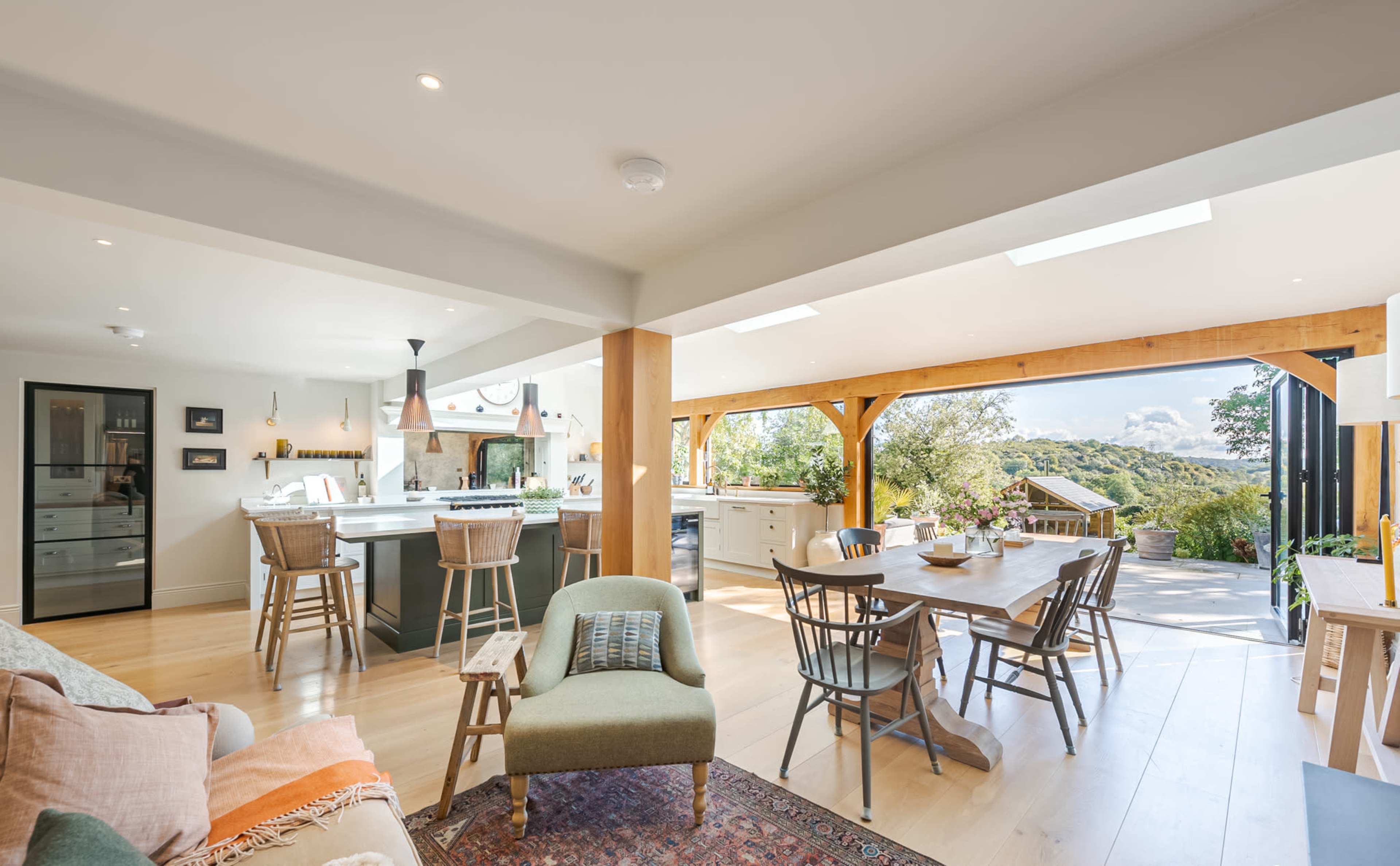 A bright, modern kitchen and dining area with a large table, wooden beams, and windows overlooking a natural landscape.